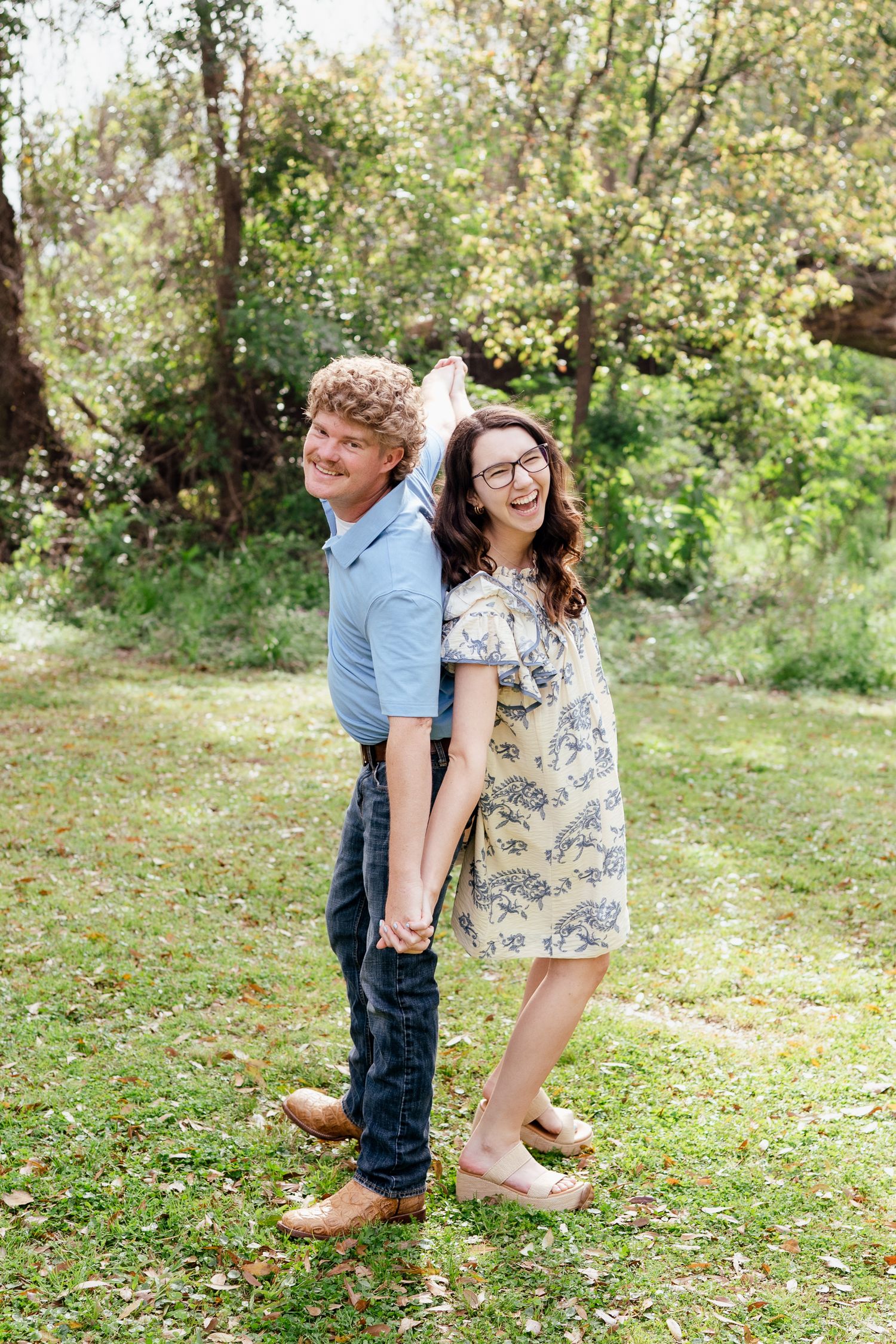 A playful couple holds hands back-to-back in a garden setting.