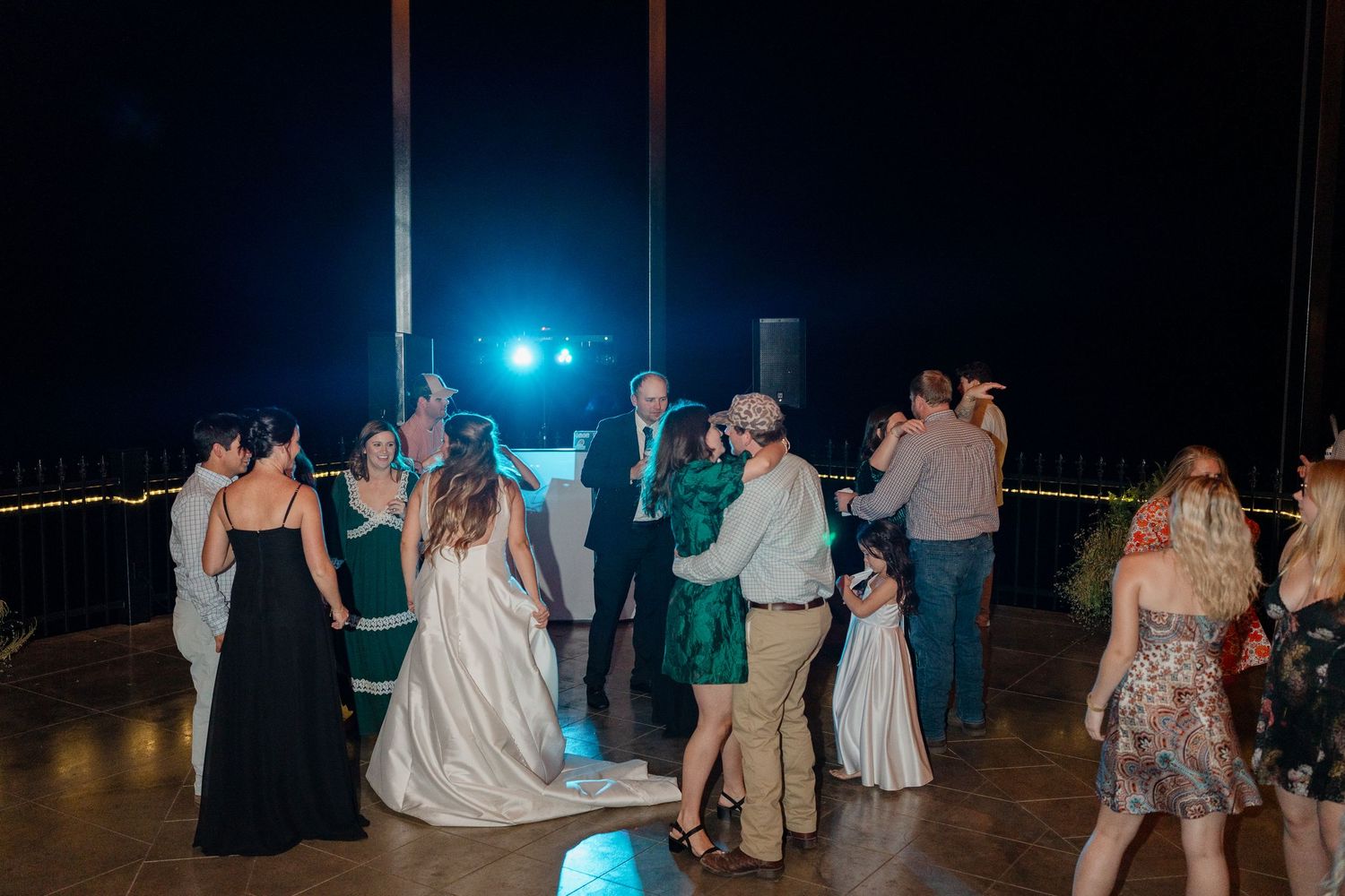 Wedding guests dance together on a dimly lit dance floor at an evening reception celebration.