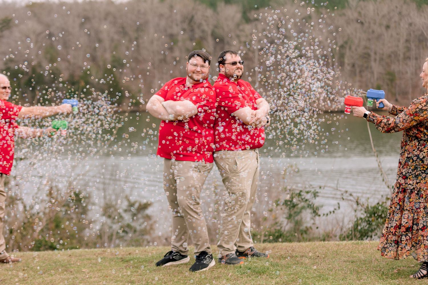 Two men in red shirts laugh as confetti is sprayed on them outdoors by a lake.
