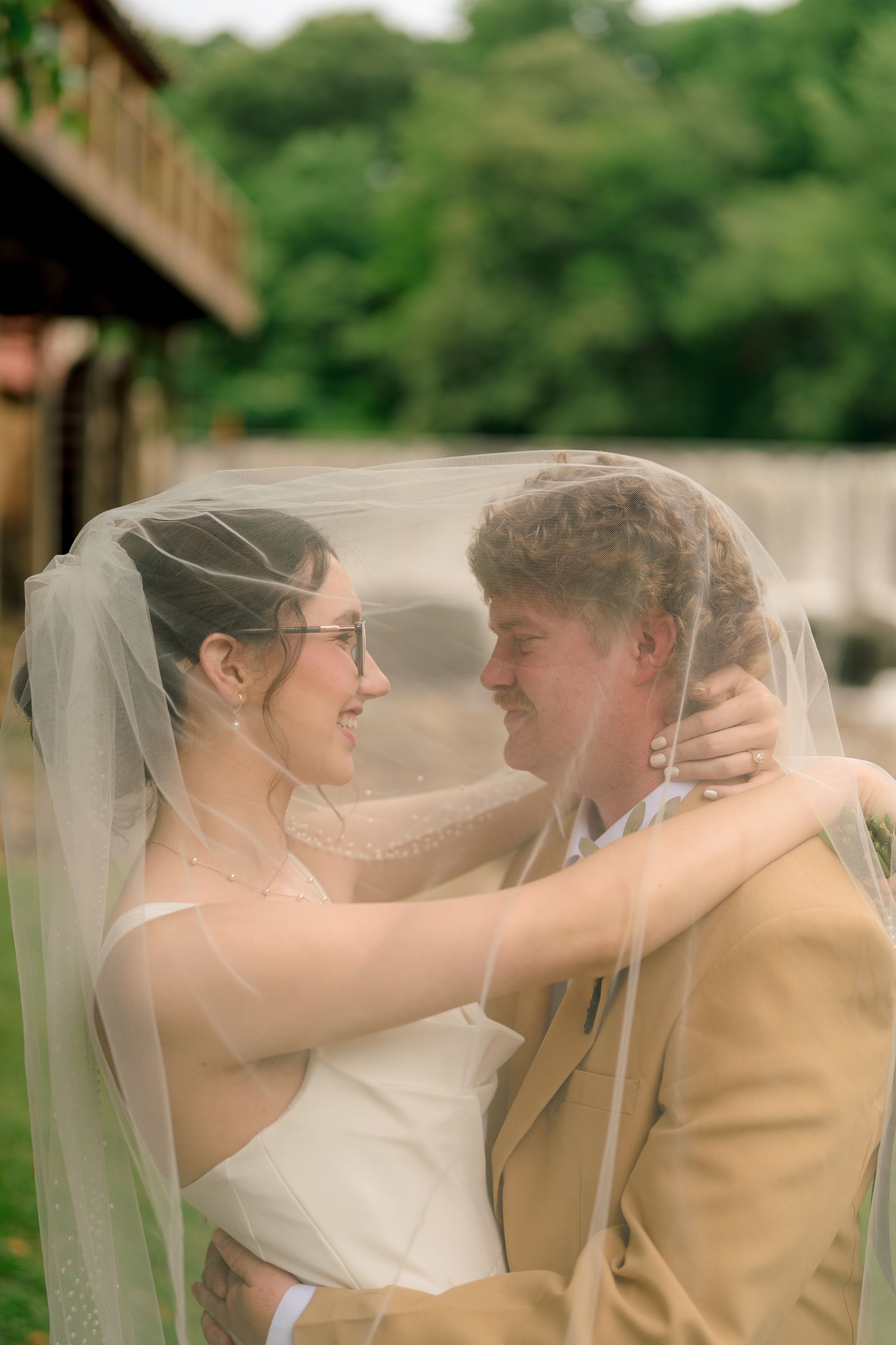 Bride and groom share romantic moment under sheer wedding veil outdoors with lush green trees behind them.