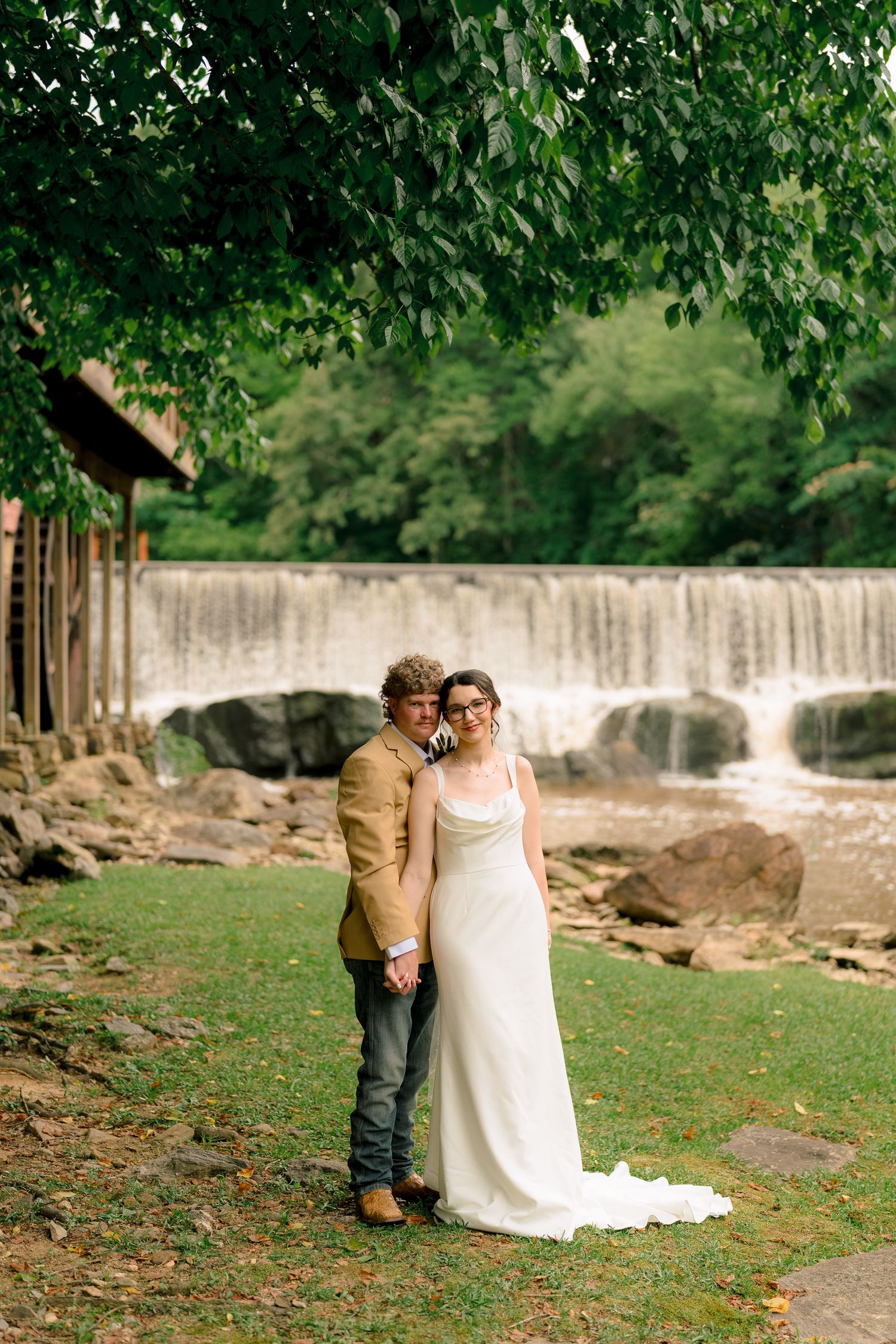 Bride and groom stand together on green lawn with scenic waterfall and lush trees in the background.