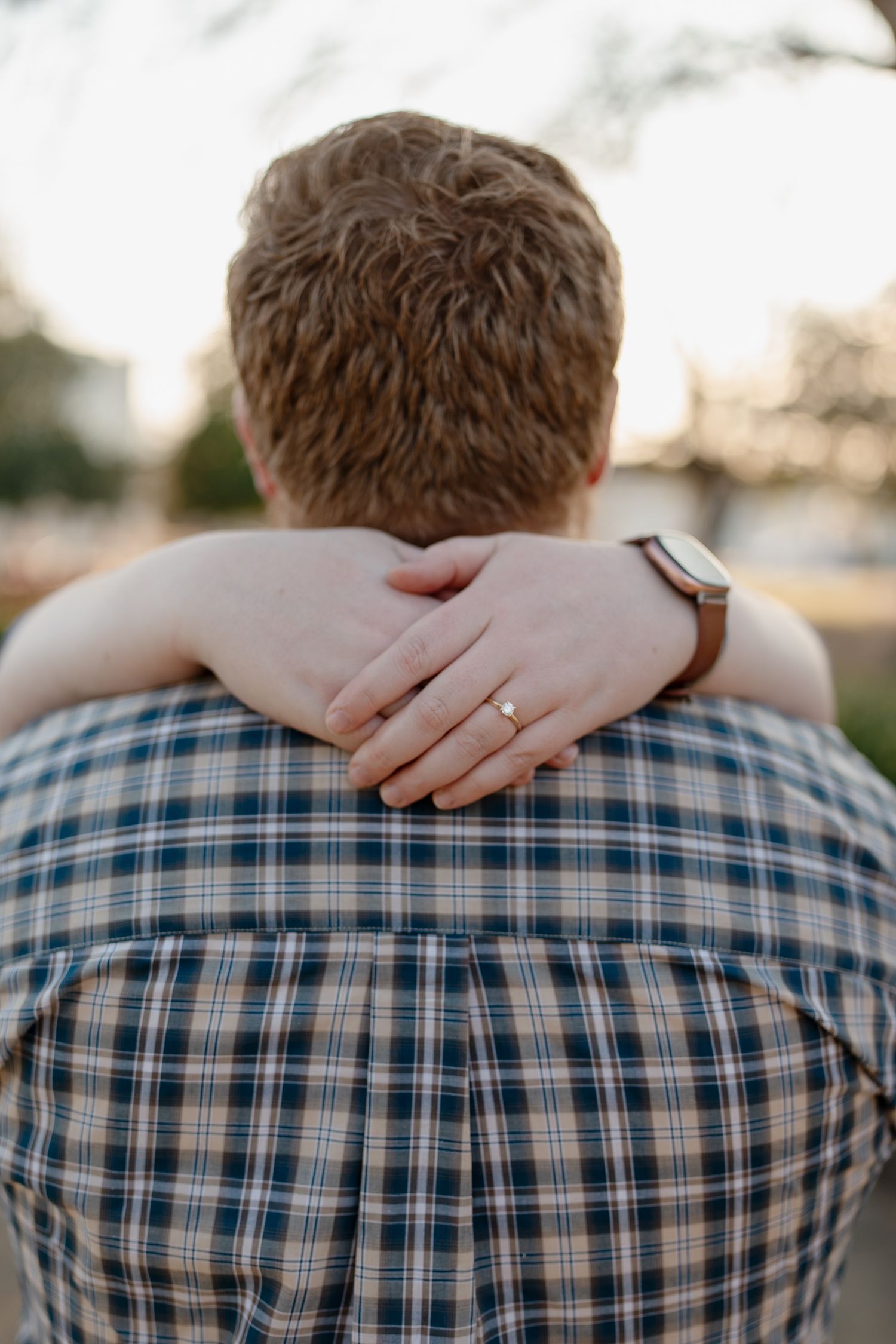 A woman's hand with an engagement ring rests on a man's shoulder wearing a plaid shirt, viewed from behind.
