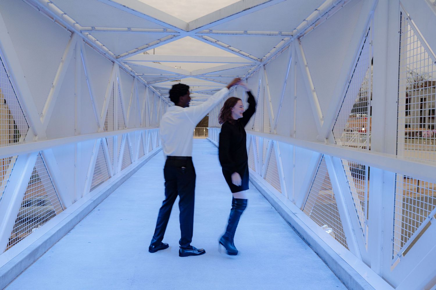 A couple dances playfully inside a bright white geometric pedestrian bridge tunnel.
