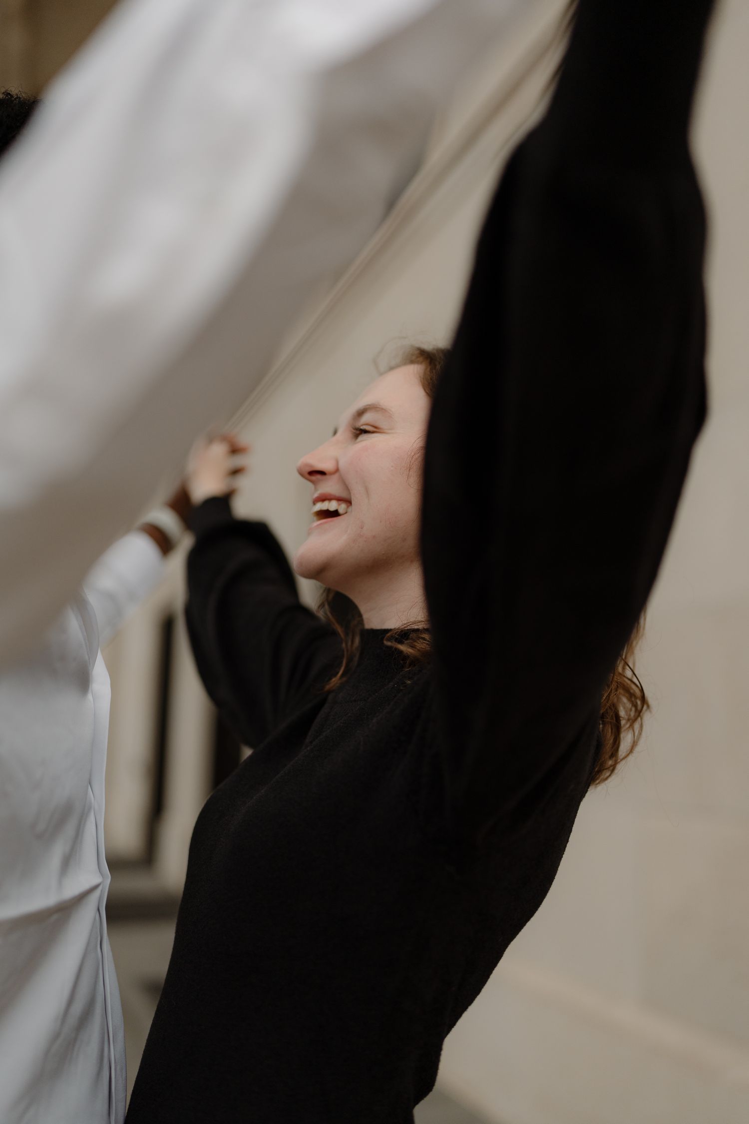 A joyful young woman in a black sweater laughs while holding up a flowing white fabric outdoors.
