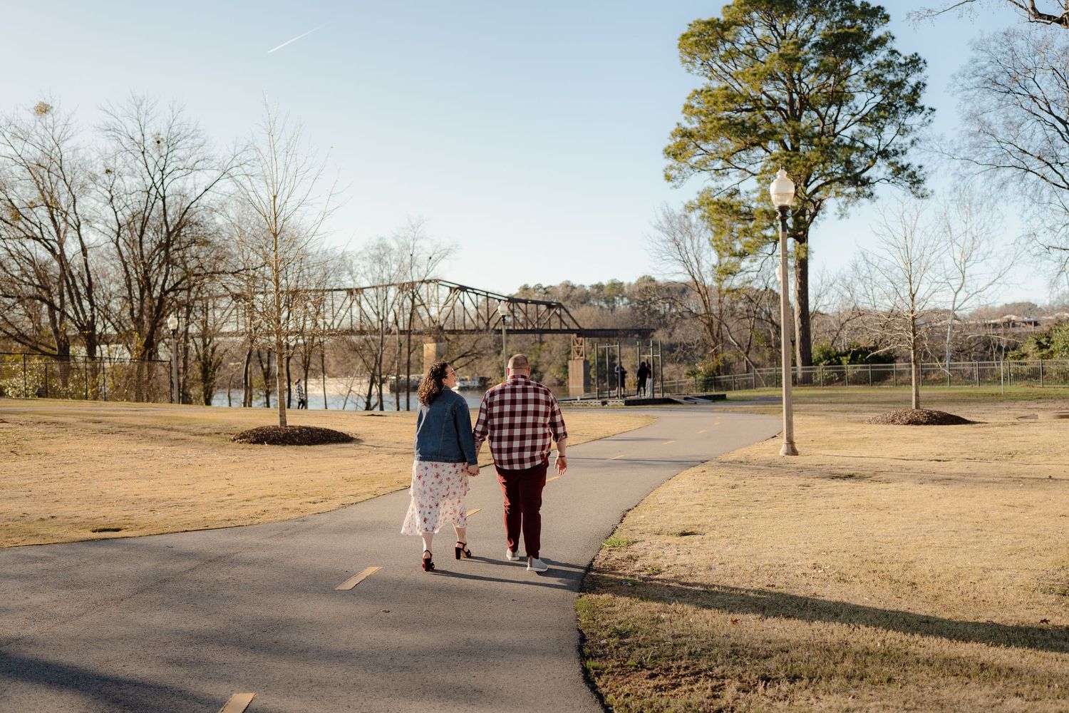 A couple walks hand in hand along a path with a metal bridge and bare trees in the background.