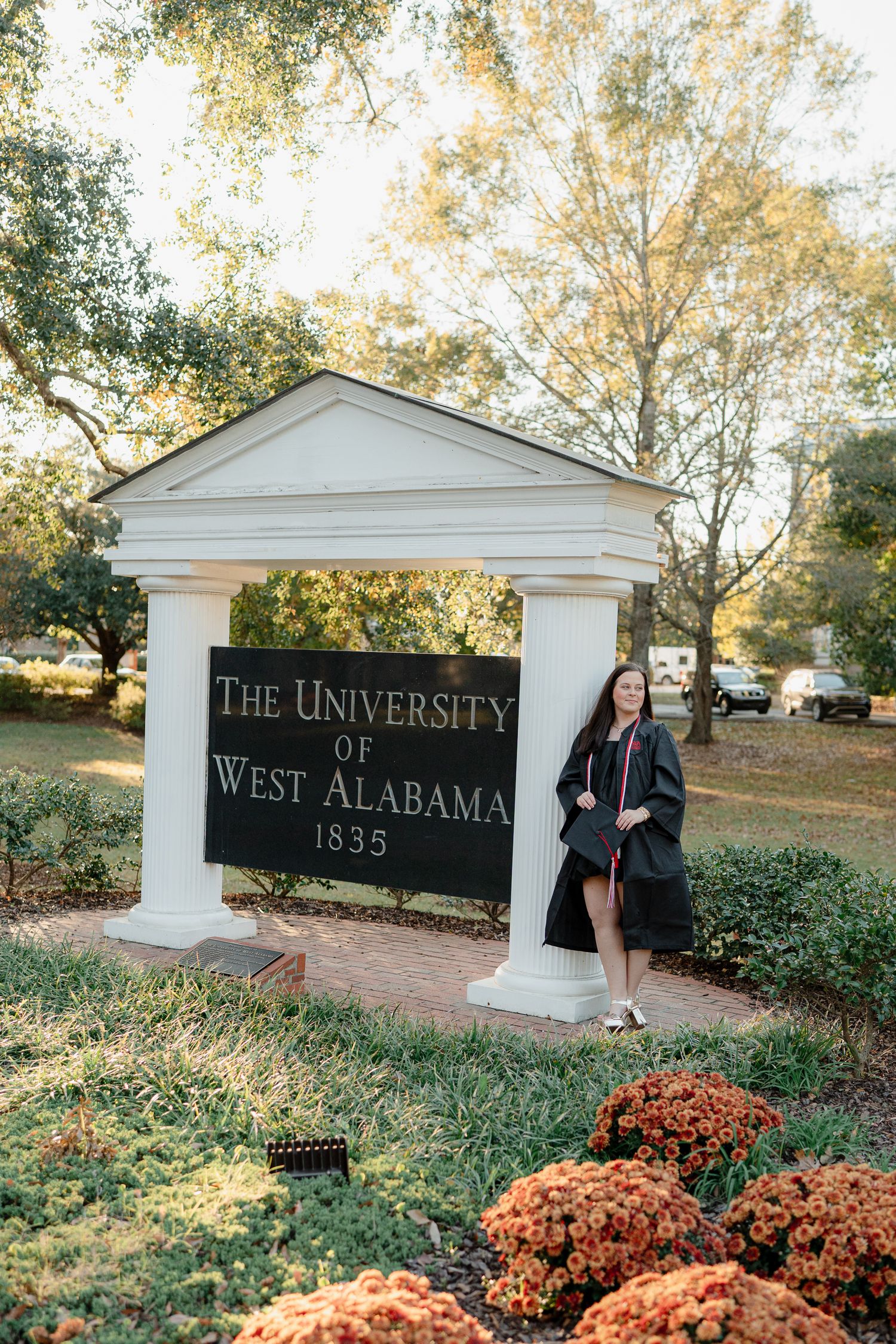 A graduate poses by the University of West Alabama entrance sign surrounded by fall mums.