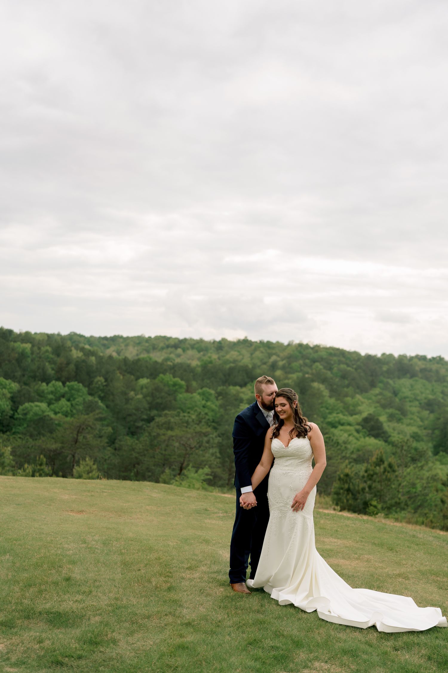 Bride and groom embrace on a green hillside with lush forest backdrop on their wedding day.