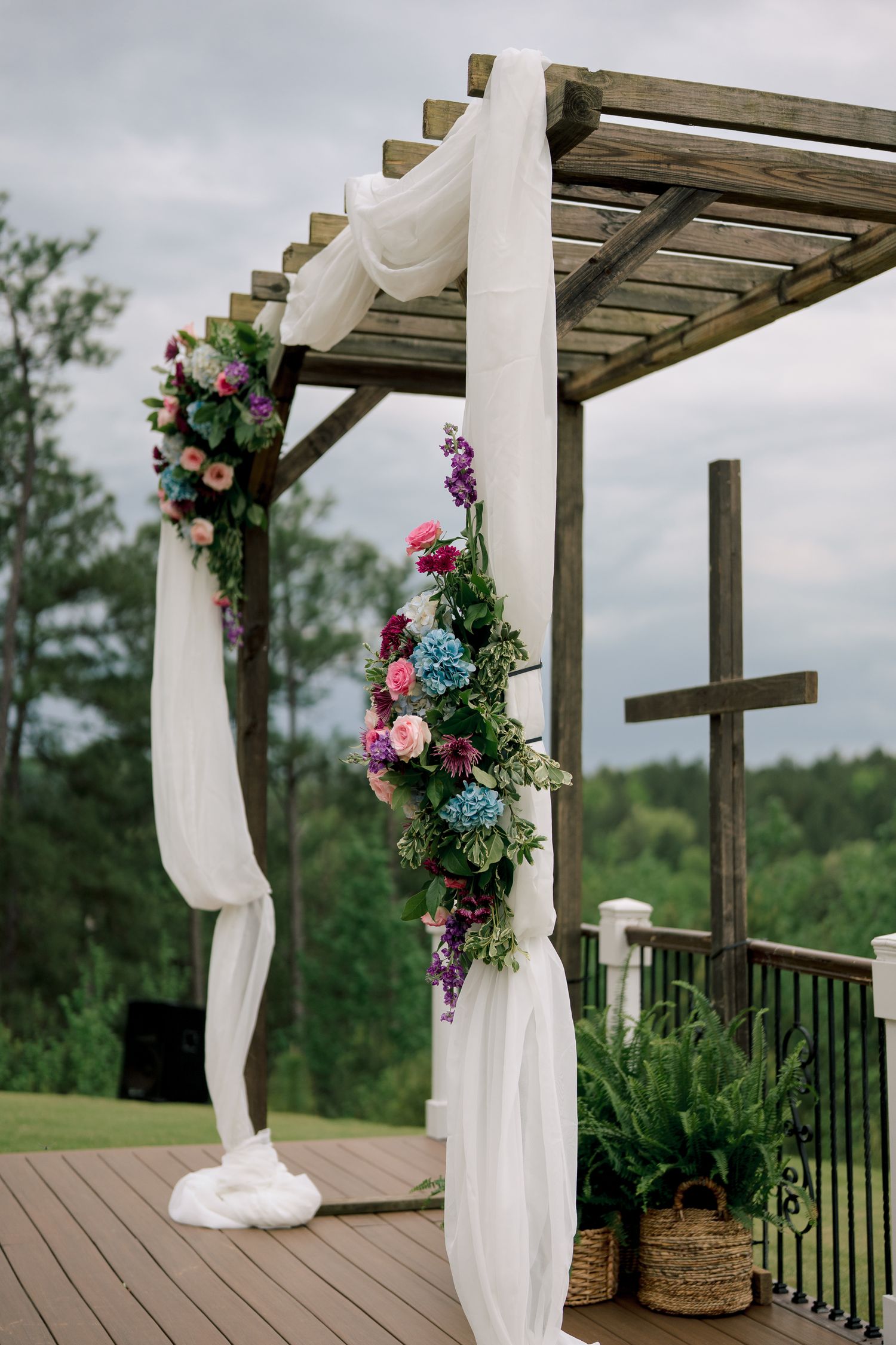 Wooden wedding pergola decorated with white draping and colorful floral arrangements on an outdoor deck.