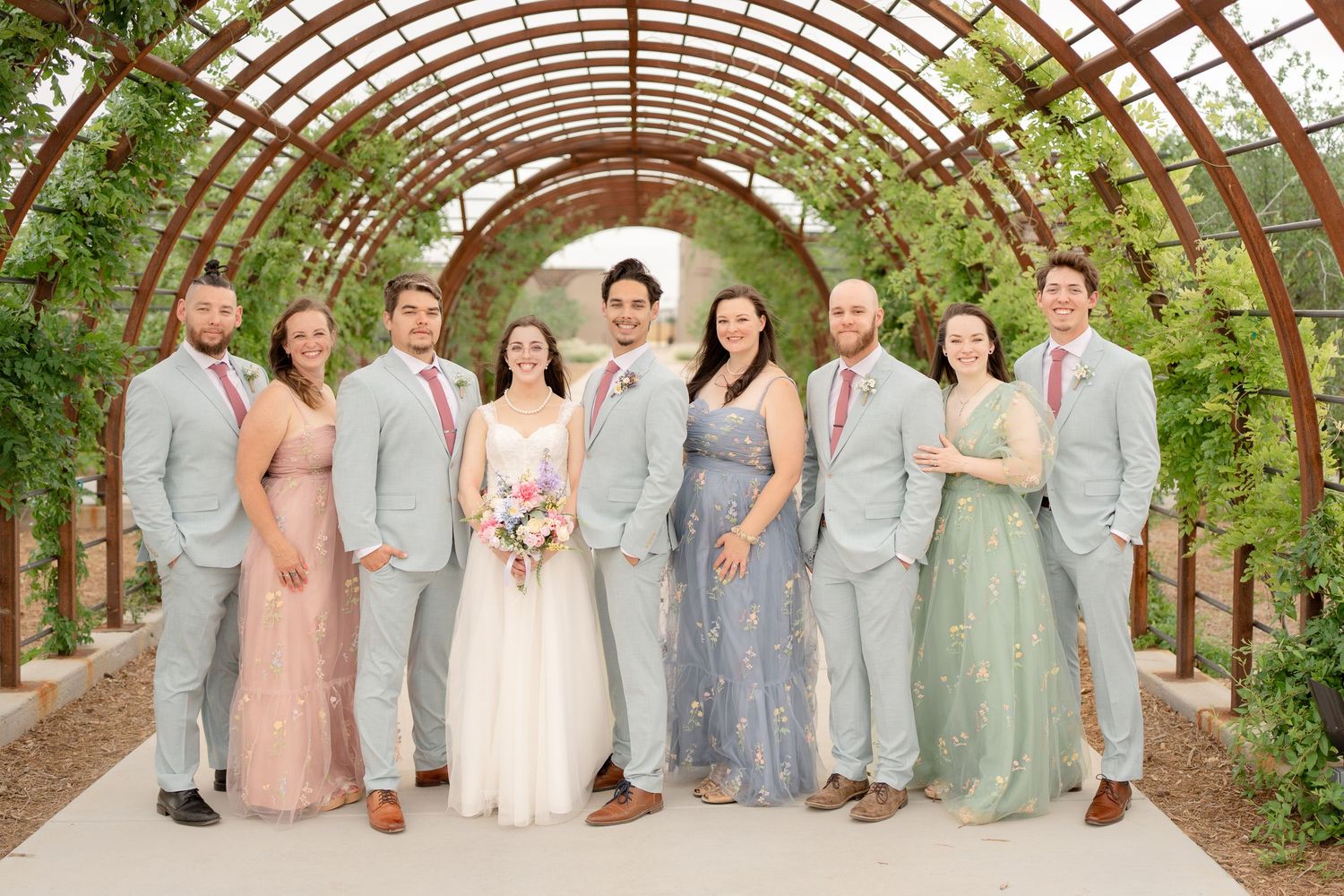 Bridal party in pastel dresses and gray suits stand together under an arched garden trellis.