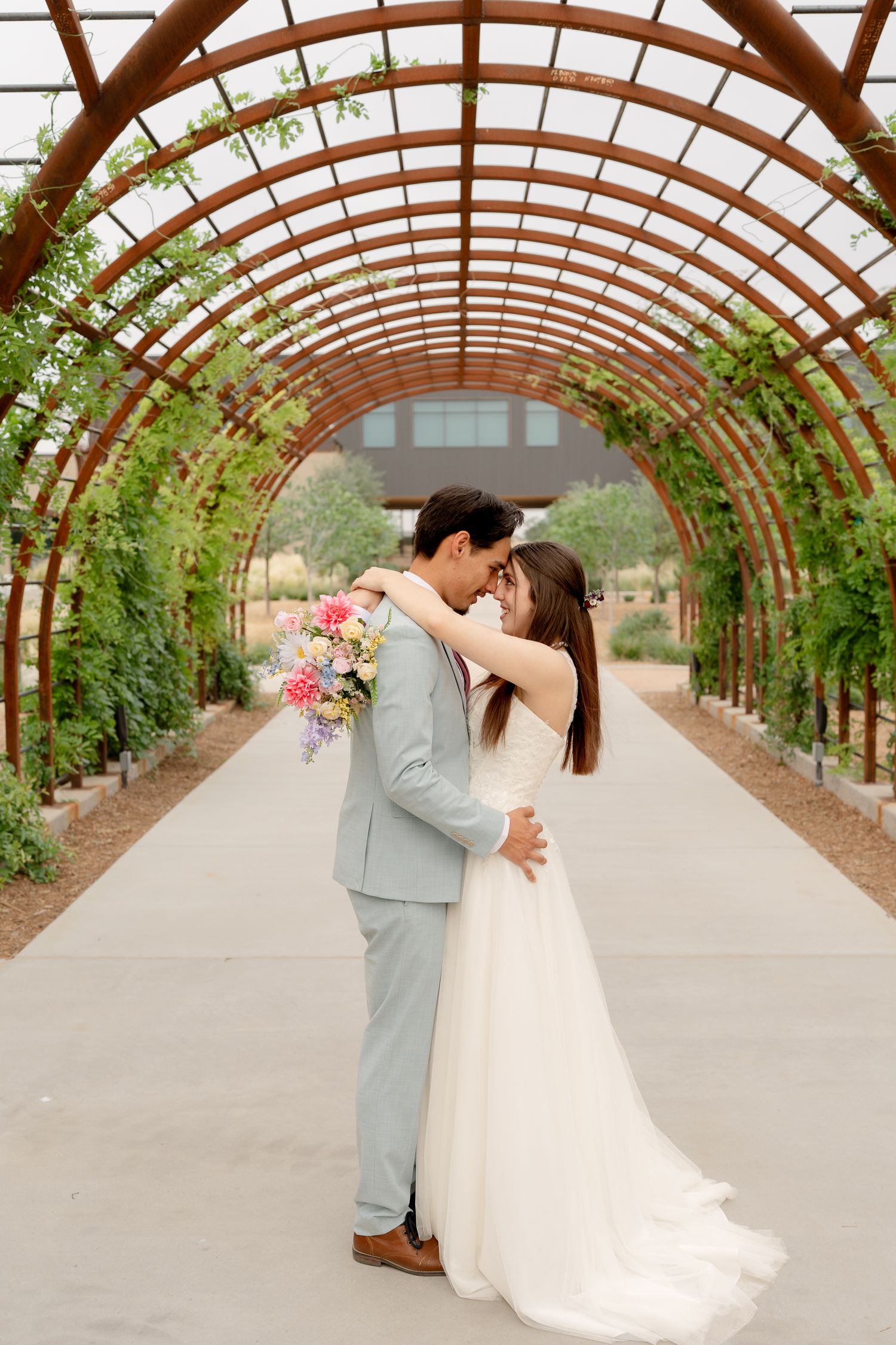 A romantic wedding moment captured under a curved wooden pergola with climbing vines and natural light.