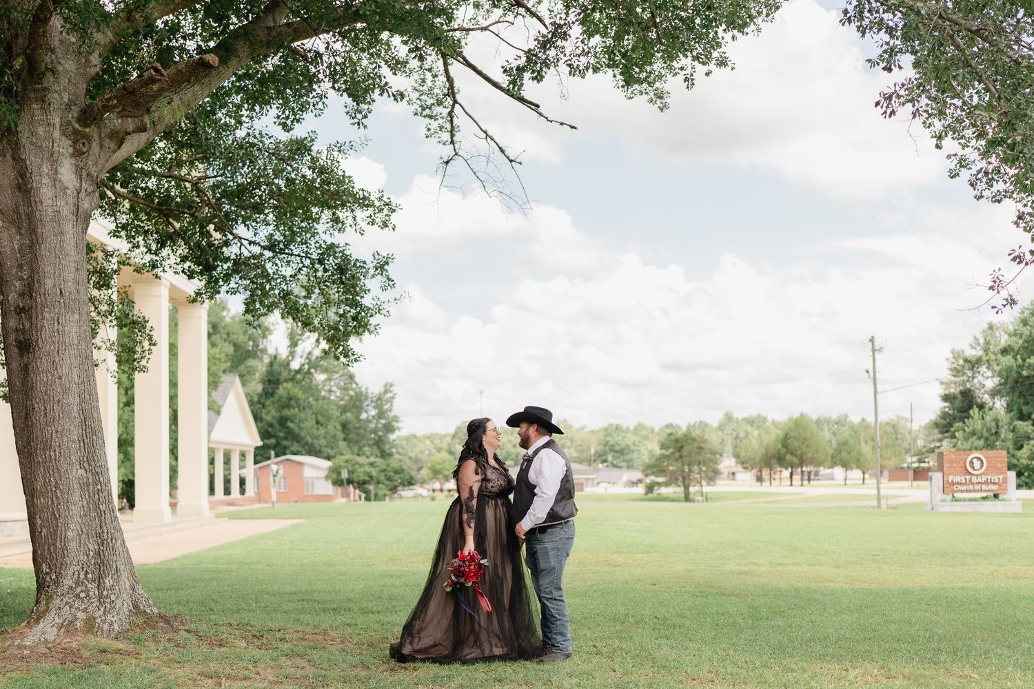 A couple in western attire shares romantic moments outdoors under trees during a wedding photoshoot.