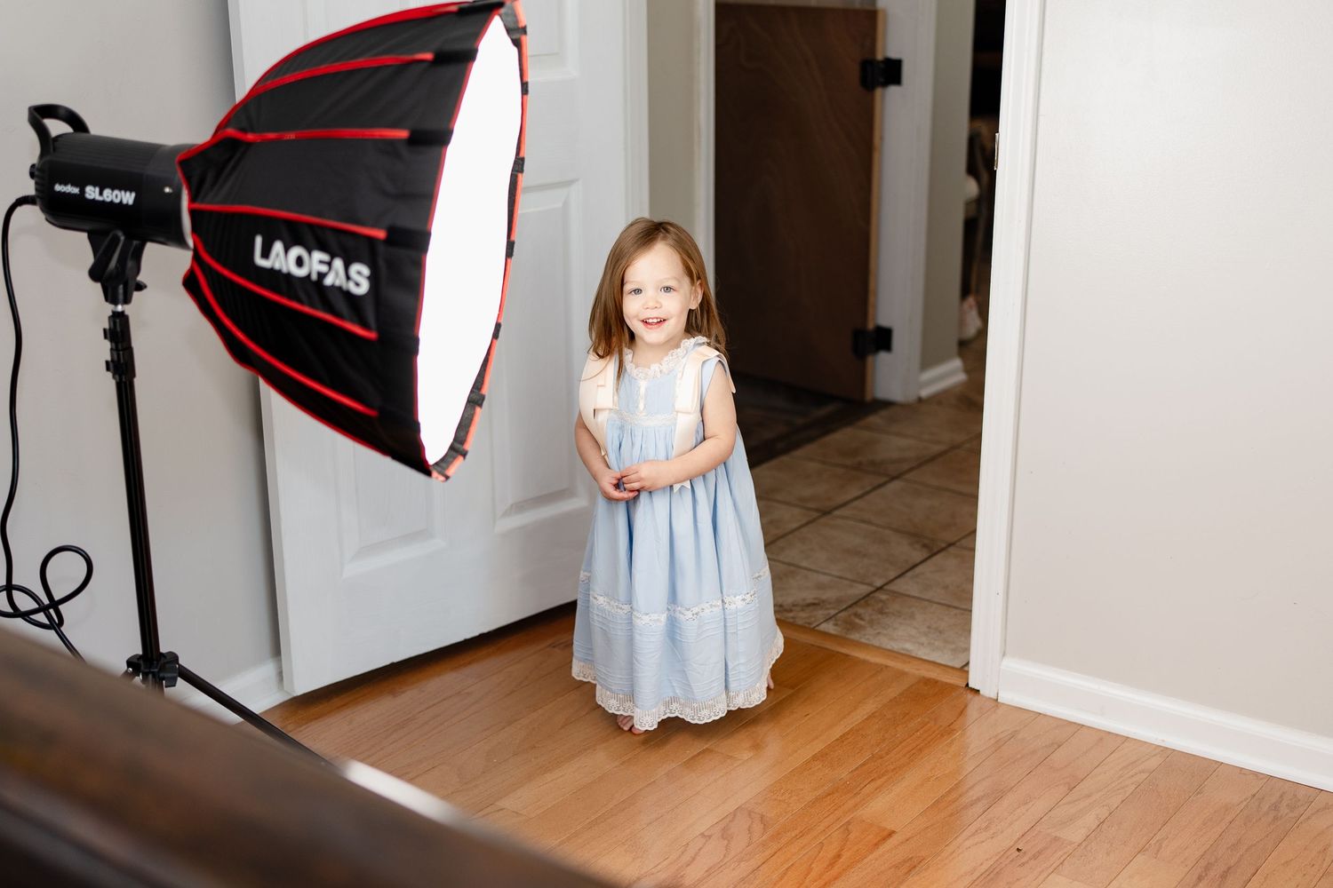 Child in blue dress standing near photography equipment during indoor photo session.