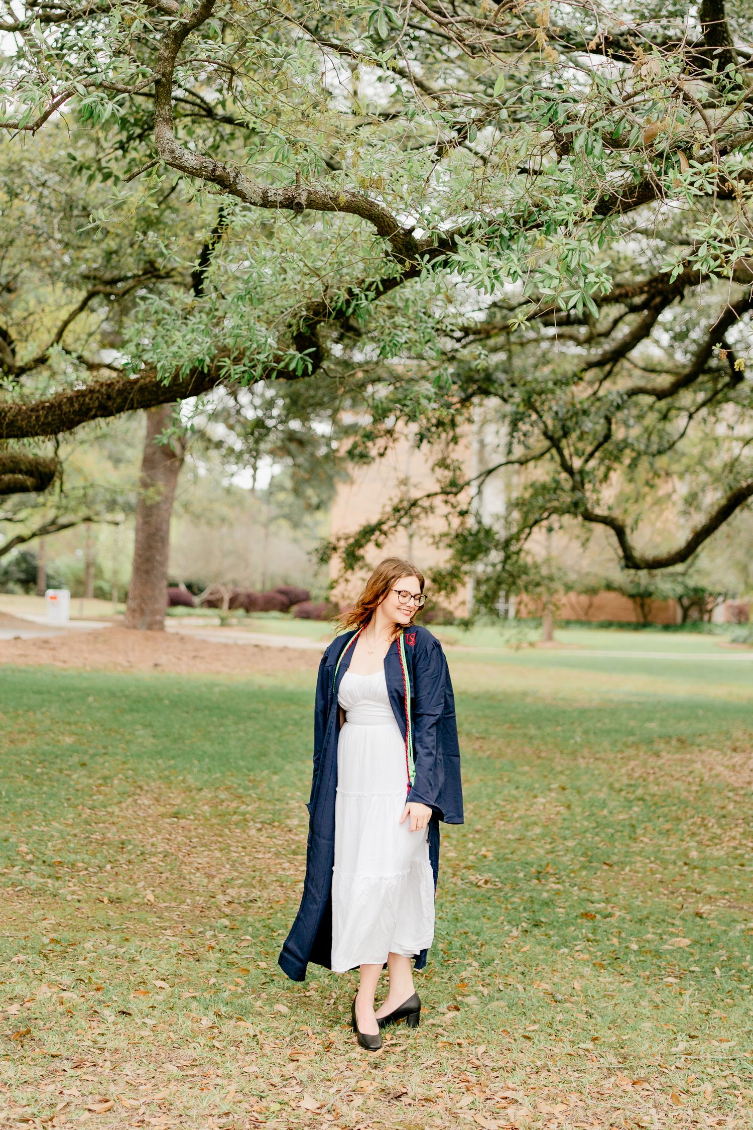 Graduate in navy gown poses under sprawling oak trees on campus lawn.