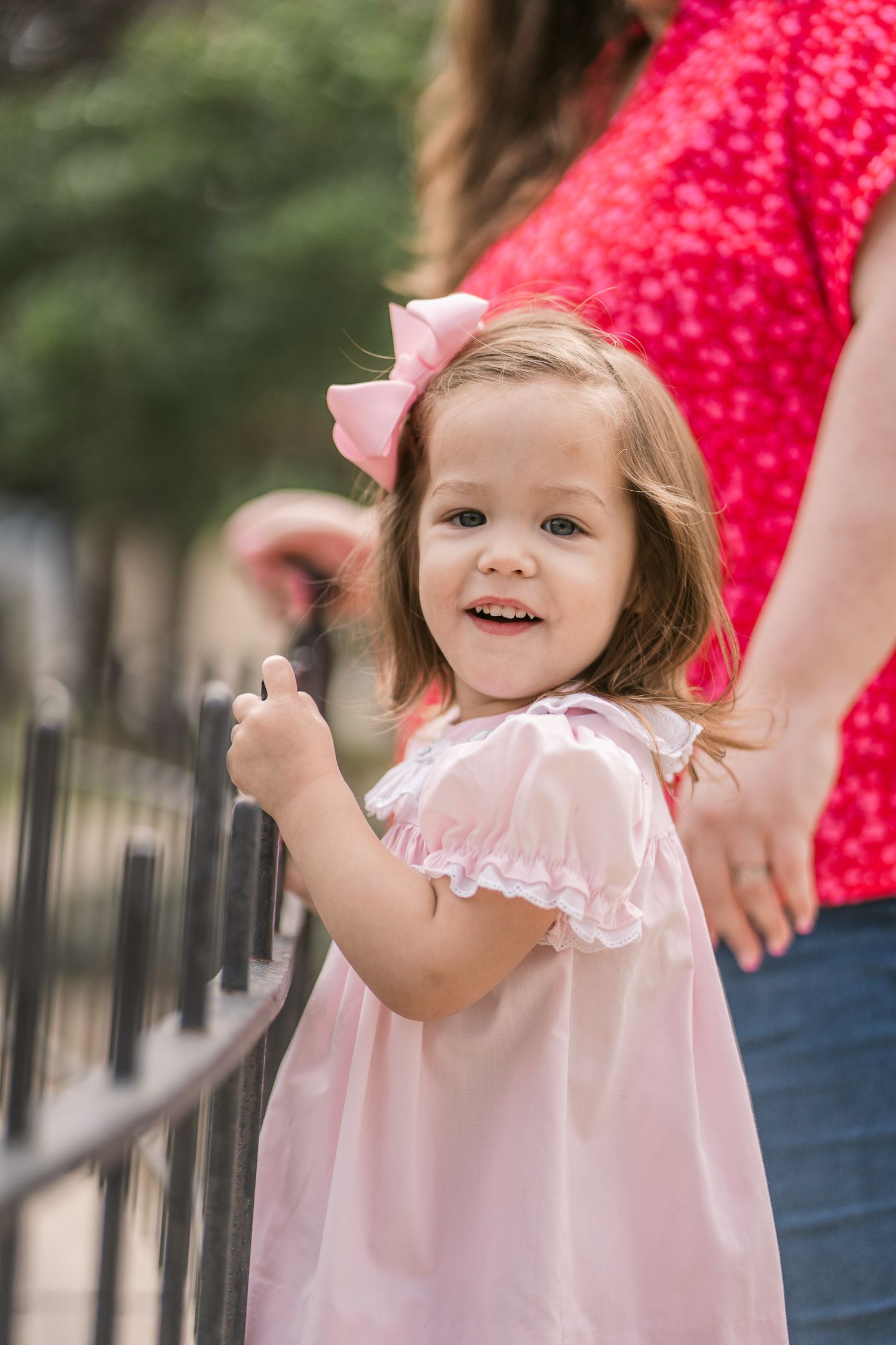 Small child wearing pink dress with bow smiles while holding onto railing with adult in red in background.