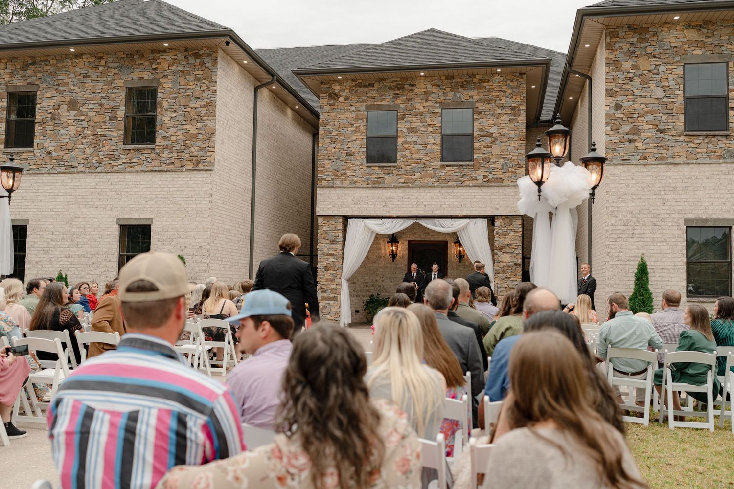 Wedding guests gather outside a stone facade venue for an outdoor ceremony with white chairs set up.