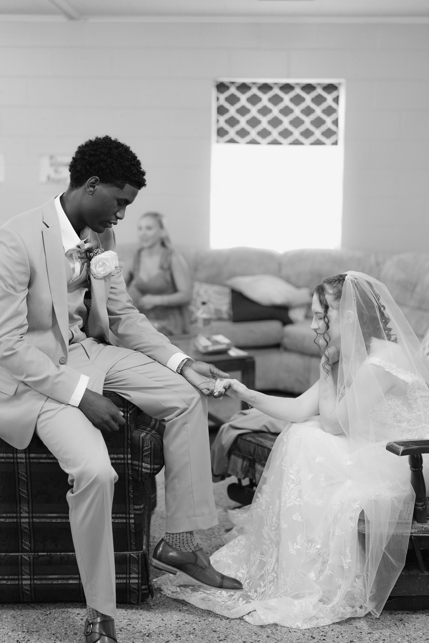 Black and white photo of groom in tan suit holding bride's hand while seated before the wedding ceremony.