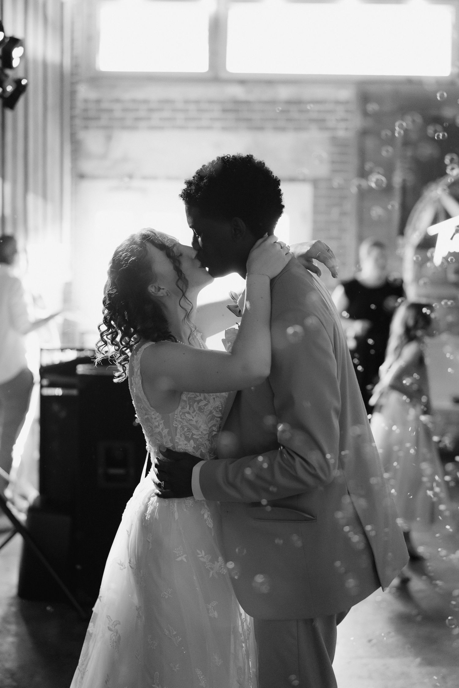 Interracial couple sharing first dance kiss surrounded by bubbles at wedding reception, black and white.