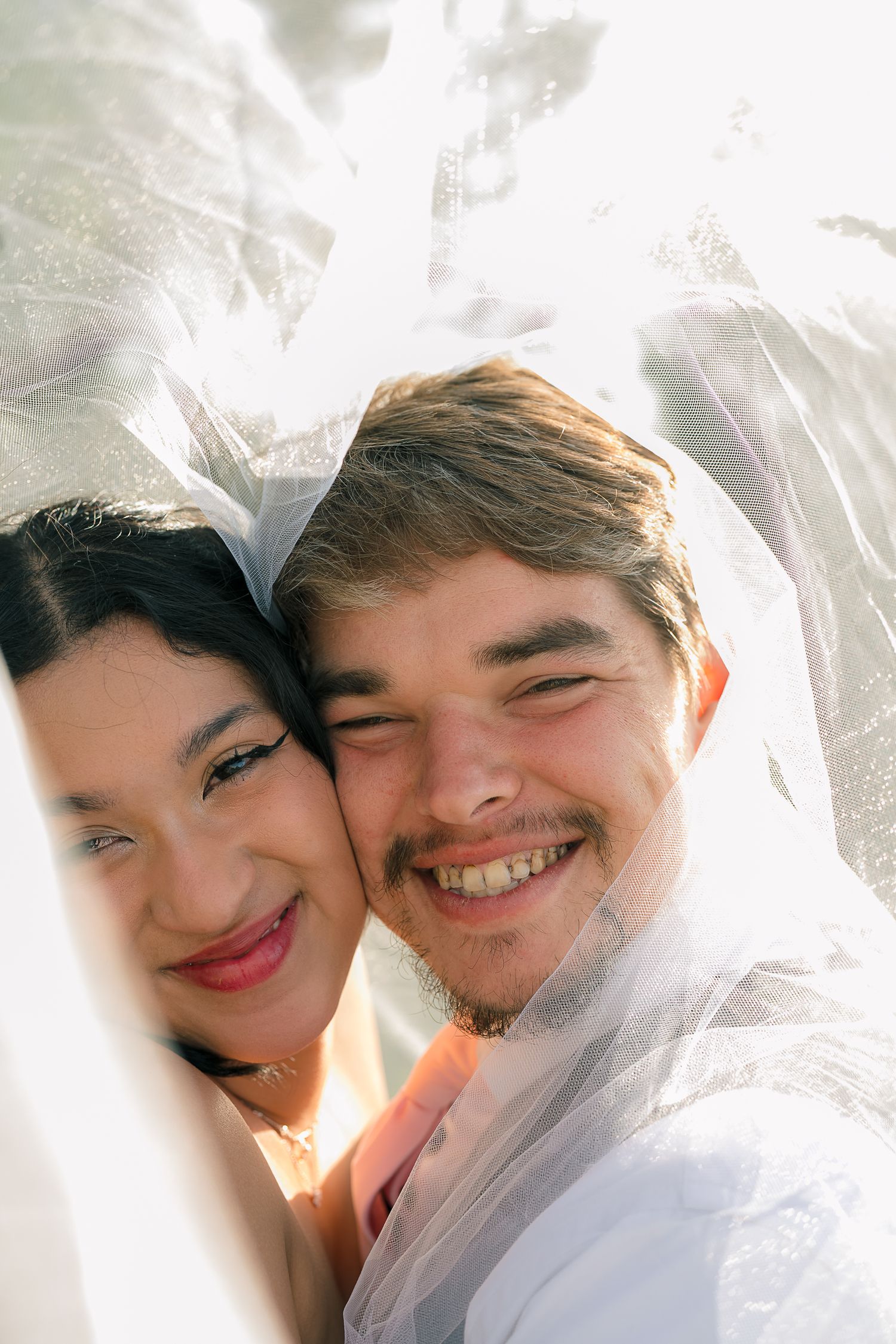 Joyful bride and groom smiling together under a sheer wedding veil in a bright outdoor setting.
