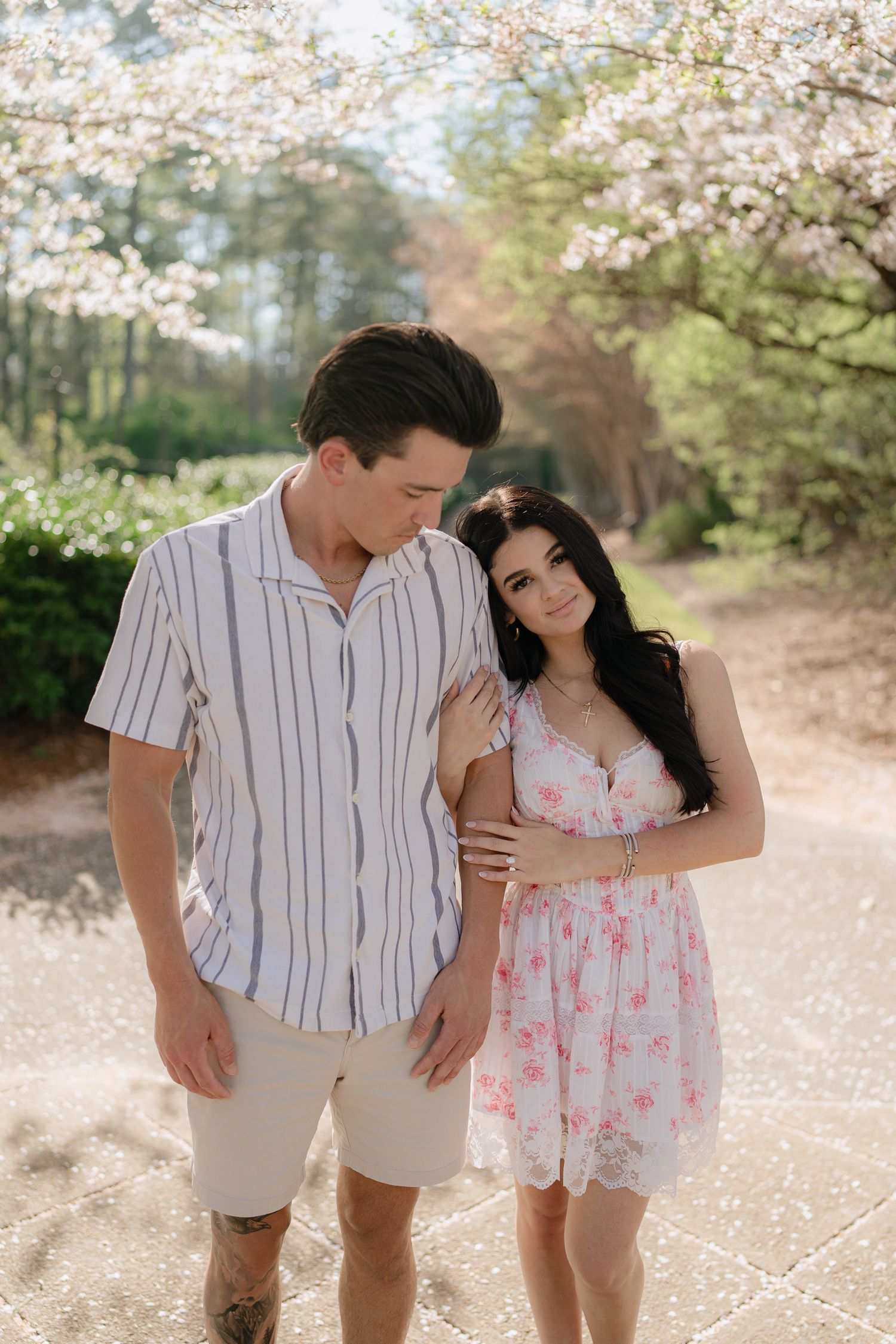 A couple walks together under blooming cherry blossom trees, woman in floral dress smiling at camera.