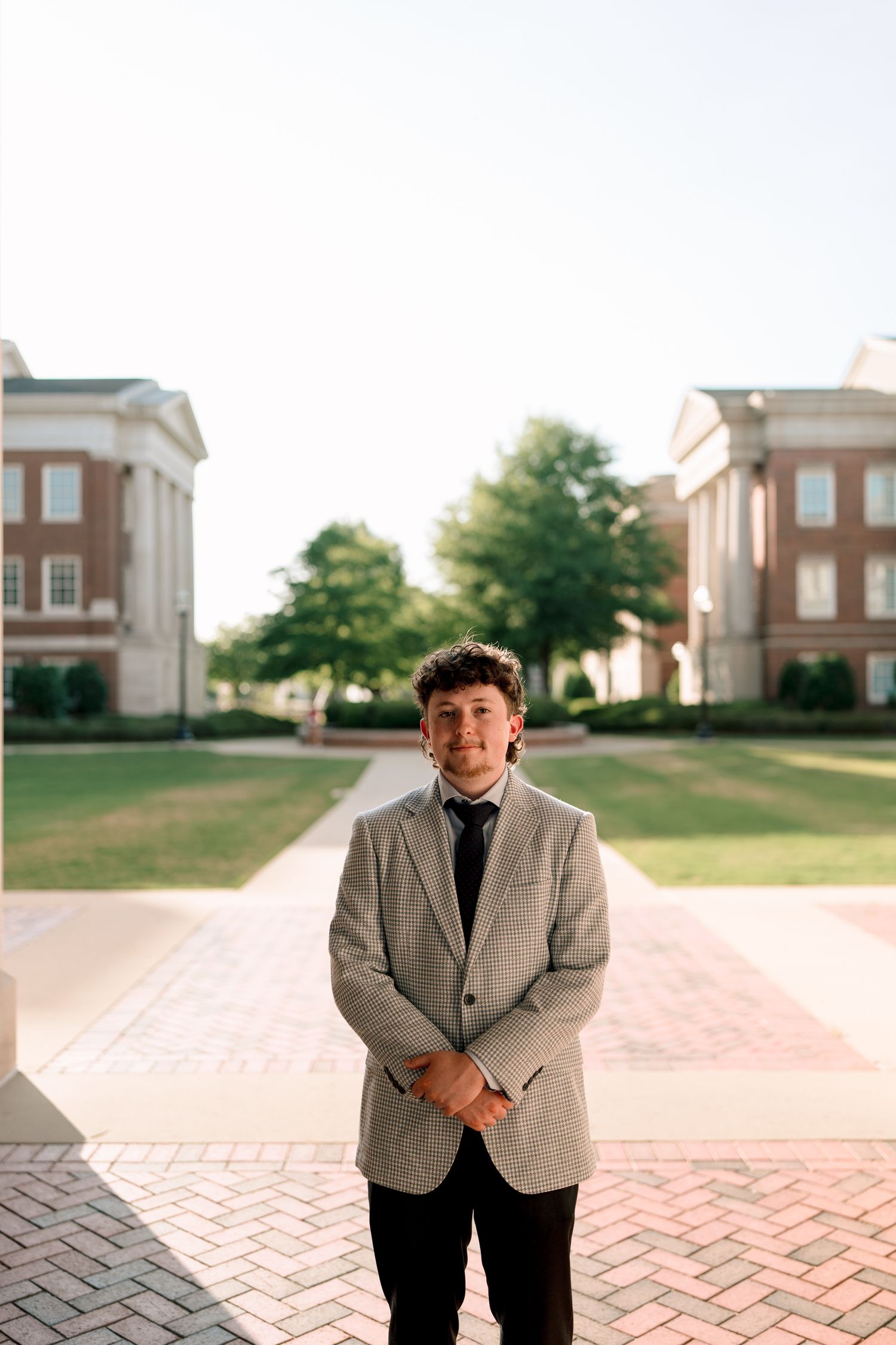 Smiling male graduate in grey blazer stands on brick plaza between two red brick campus buildings.
