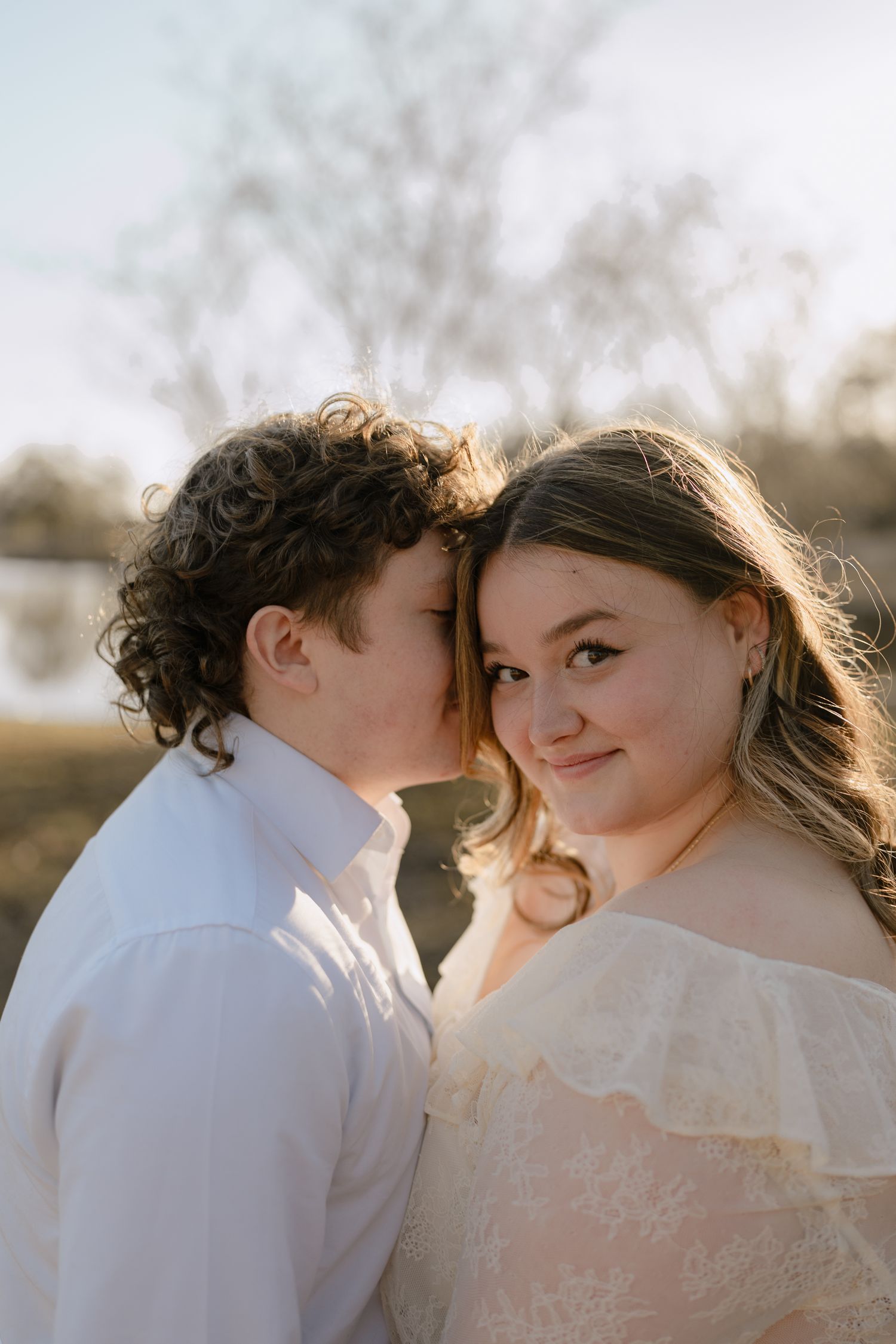 Man kissing woman on cheek as she smiles at camera in warm outdoor light.