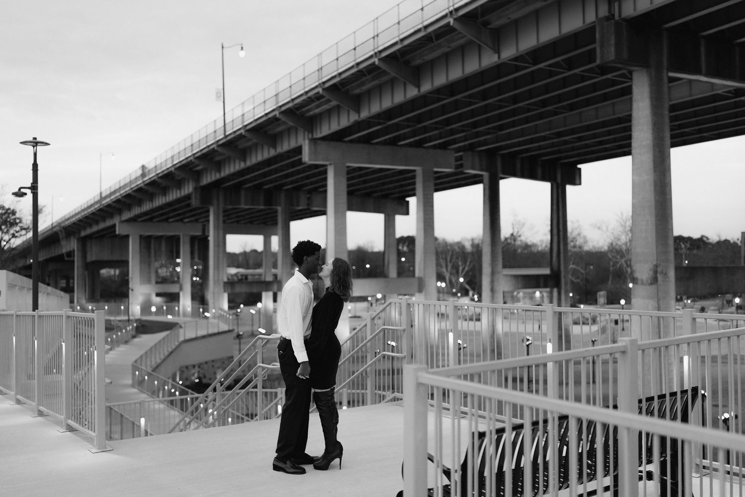 A romantic couple shares an intimate moment on a bridge walkway in a black and white urban landscape photo.