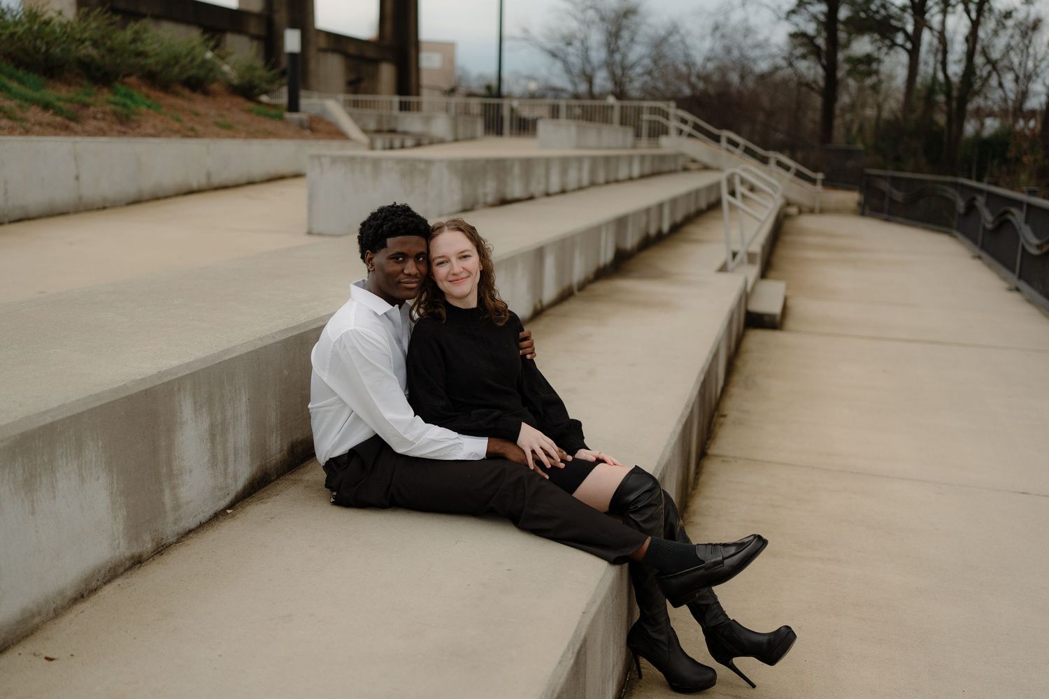 Smiling interracial couple sitting together on concrete bleacher steps, woman in black knee-high boots.