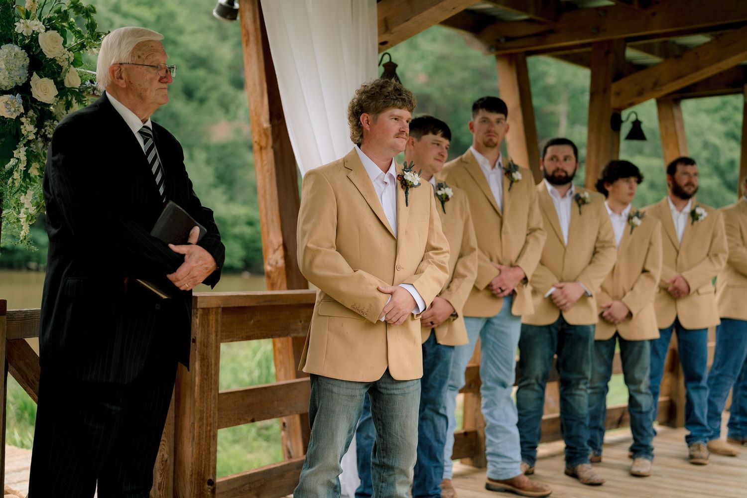 Groom in tan blazer and jeans stands with groomsmen at rustic outdoor wedding ceremony pavilion.