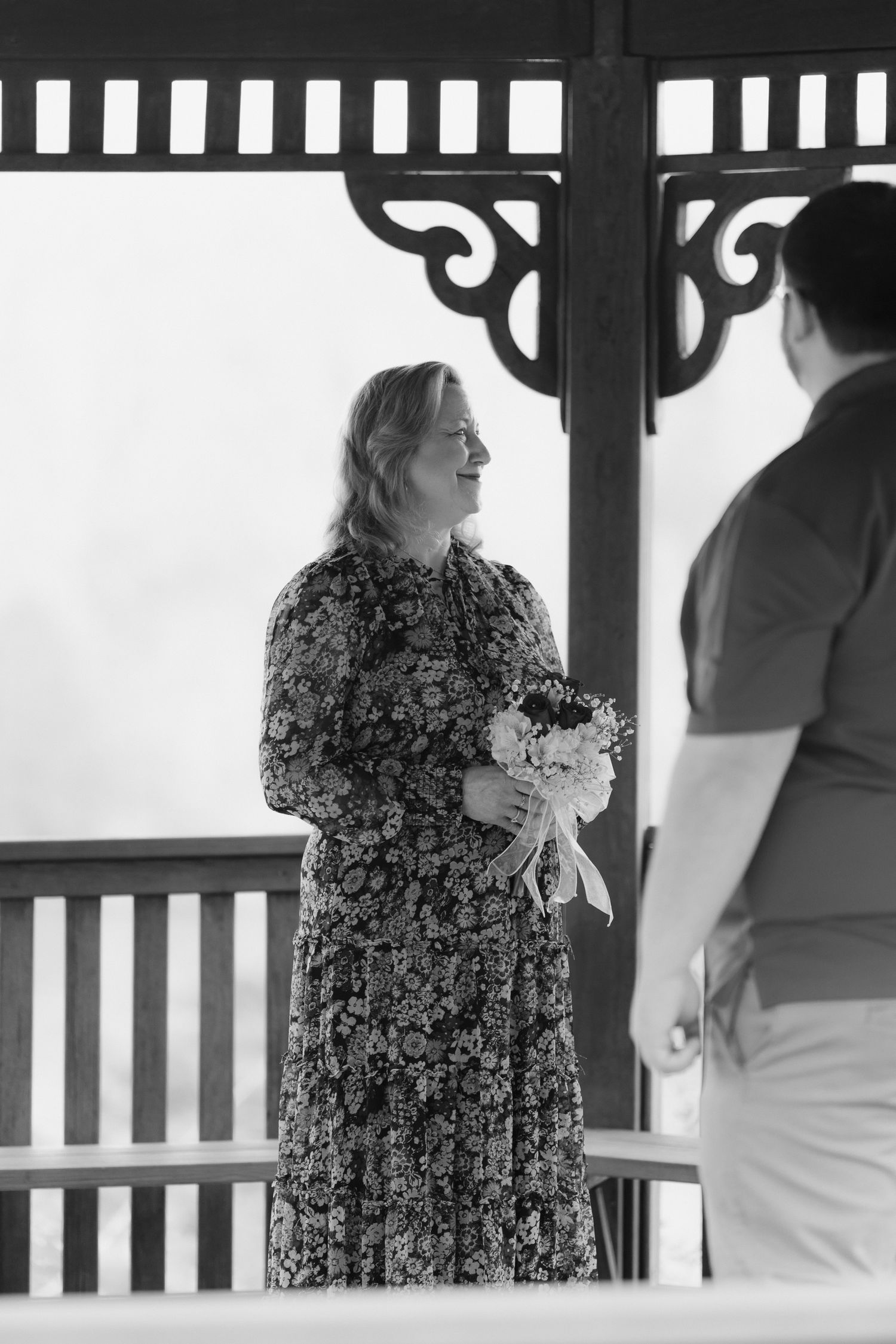 Black and white photo of a woman in a floral dress holding a bouquet inside a gazebo.