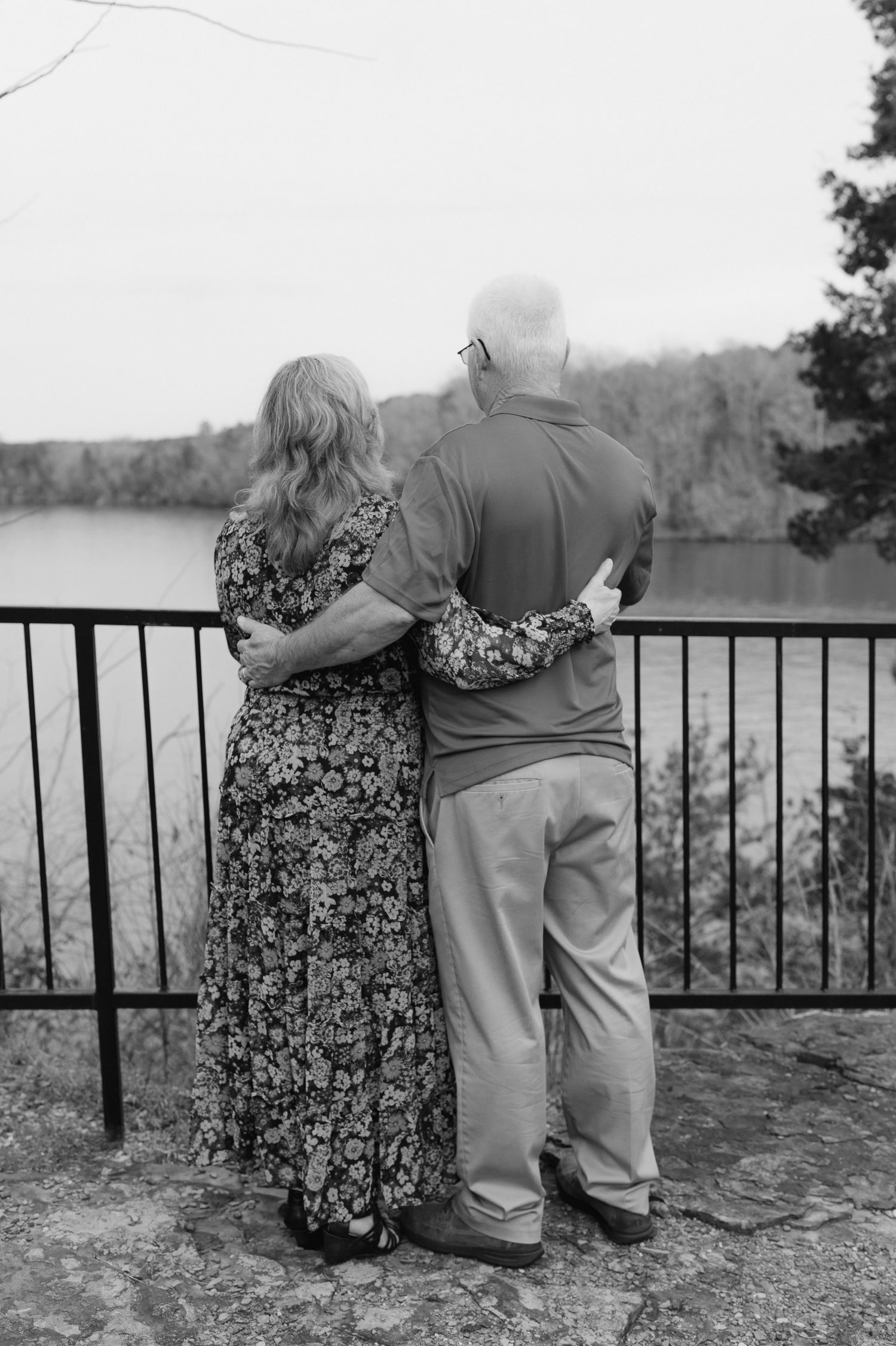 Black and white photo of a couple embracing while looking out over a lake from a railed overlook.