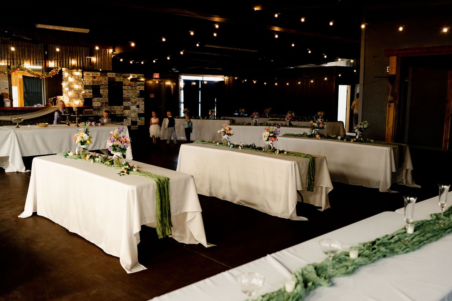 Elegant wedding reception tables decorated with white linens and greenery garlands in a dimly lit venue.