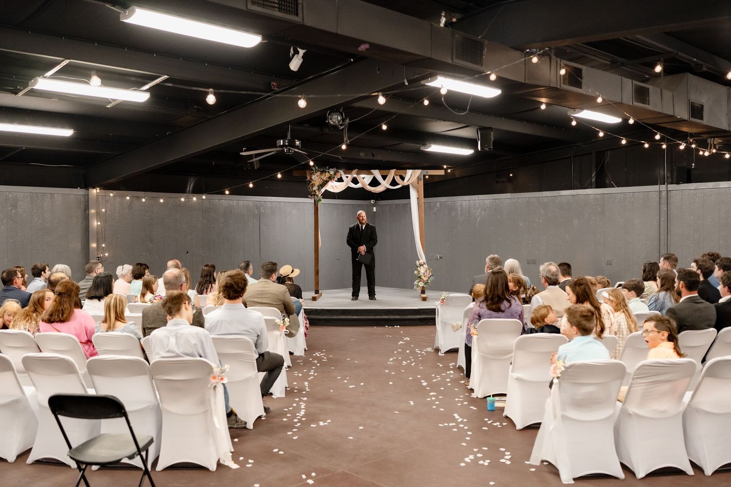Wedding ceremony setup in an industrial venue with white chairs arranged in rows under exposed ceiling beams and string lights.