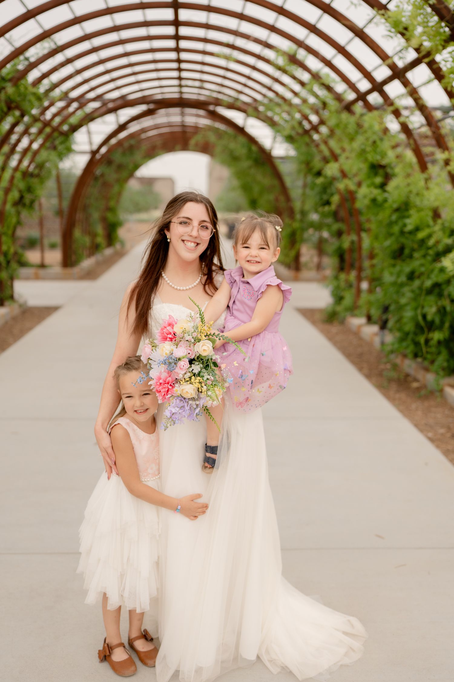 Two people in white dresses share a joyful moment together in a greenhouse setting with curved wooden beams overhead.