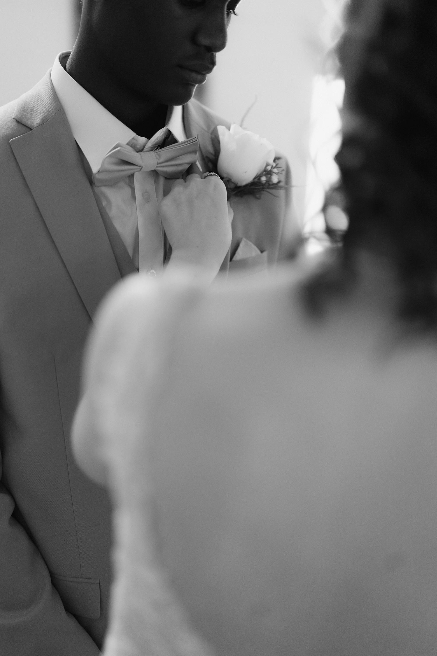 Black and white photo of bride adjusting groom's bow tie and boutonniere on wedding day.