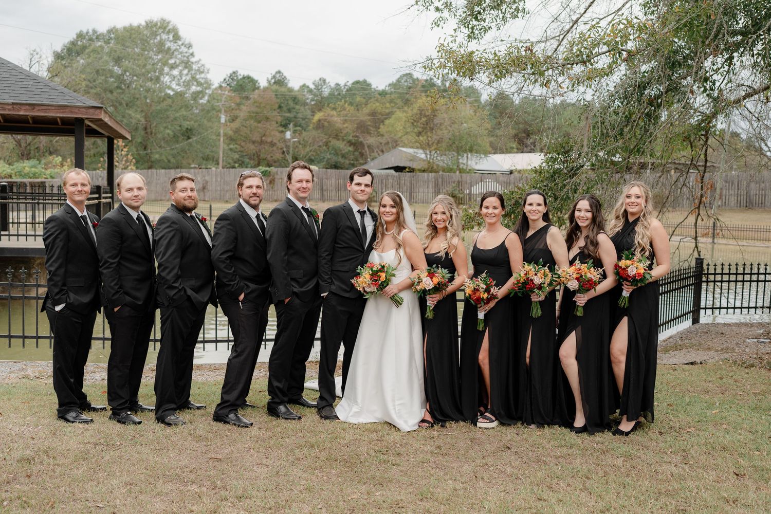 Wedding party in black attire and white gown pose together on grassy lawn for group photo.
