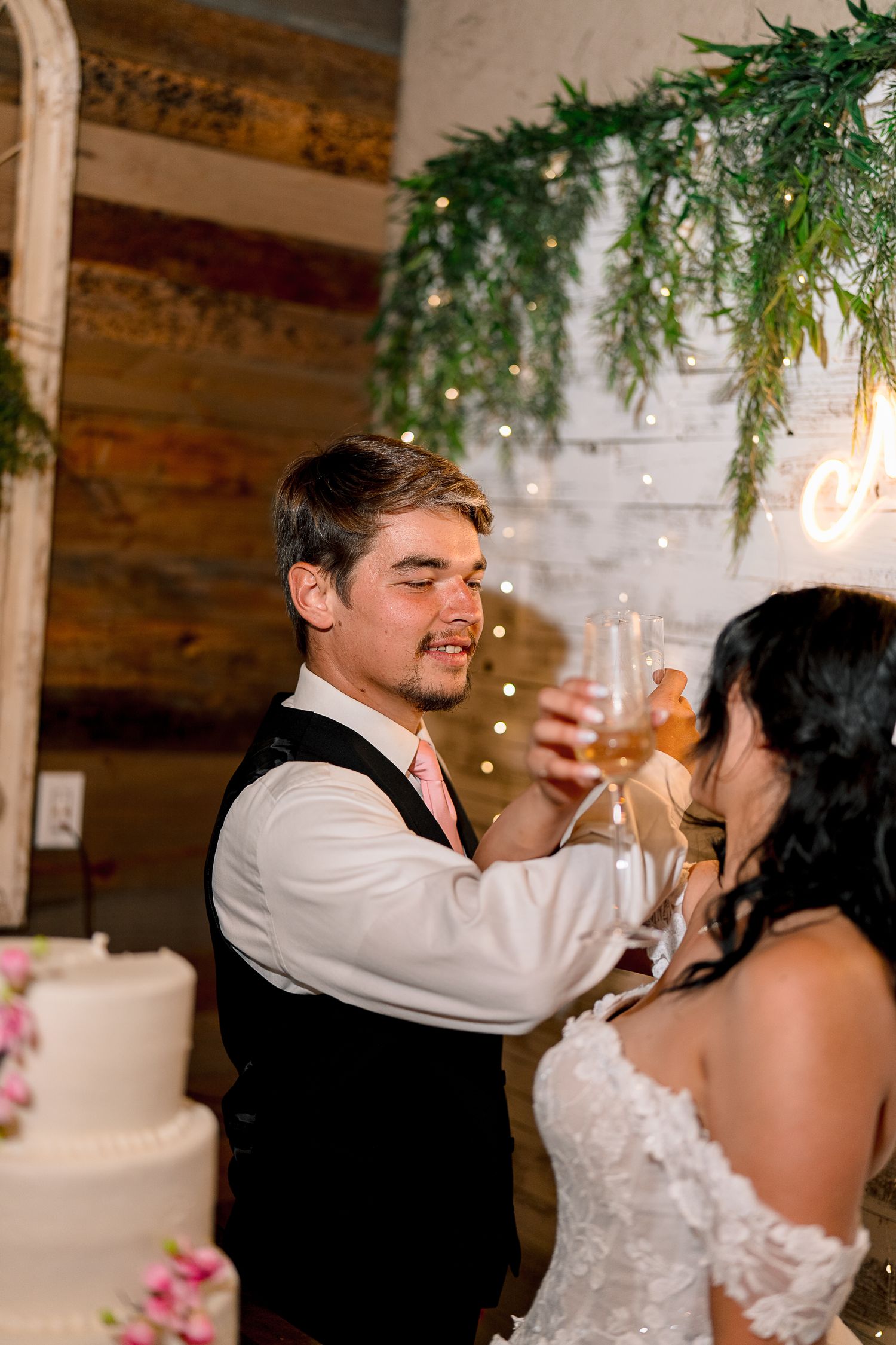 Bride and groom toast with champagne glasses near wedding cake at rustic reception venue.