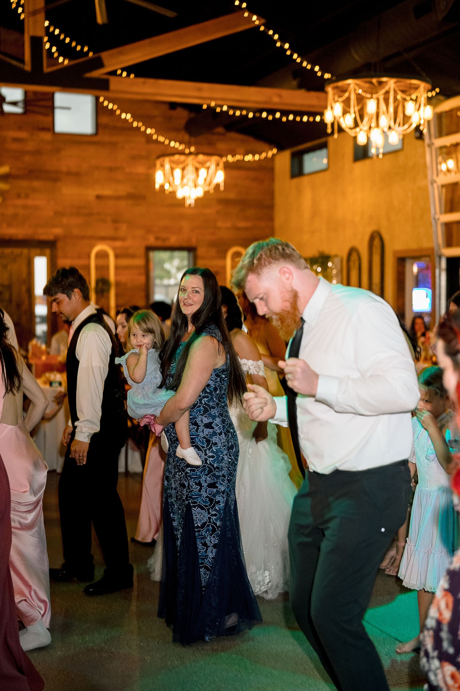 Guests dancing joyfully at a rustic wedding reception venue with chandeliers and string lights.