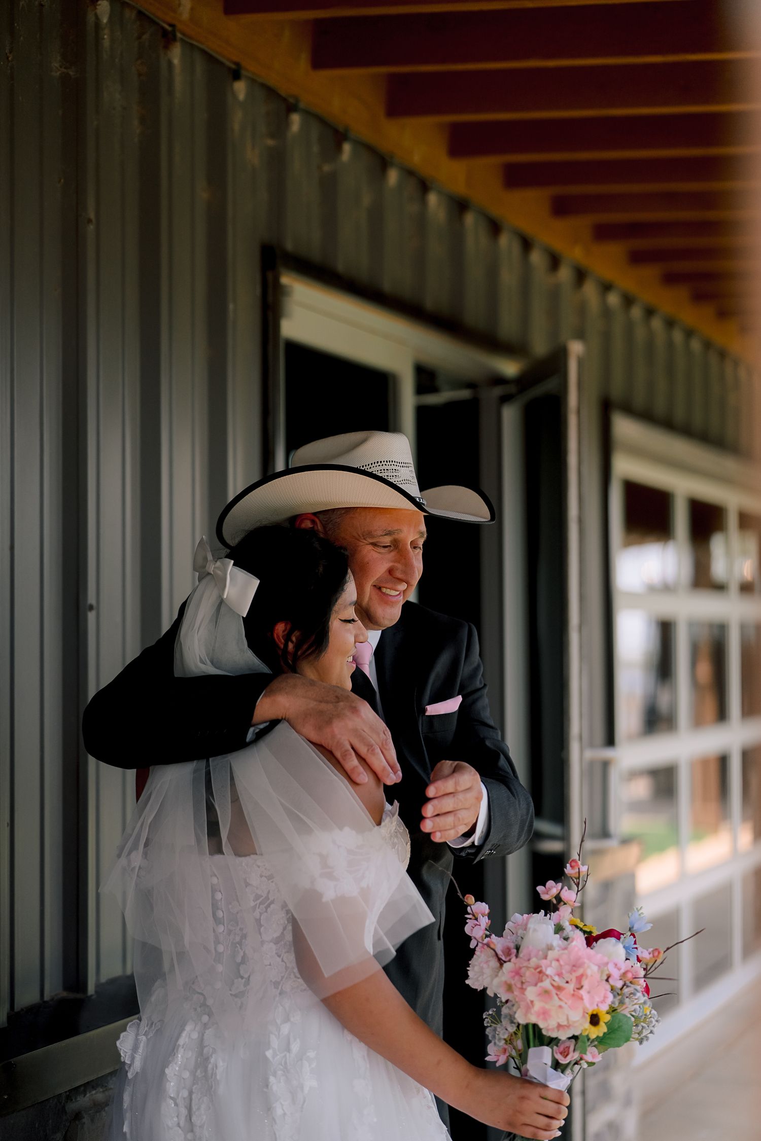 Smiling groom in cowboy hat embraces bride in white dress holding colorful bouquet outside rustic venue.