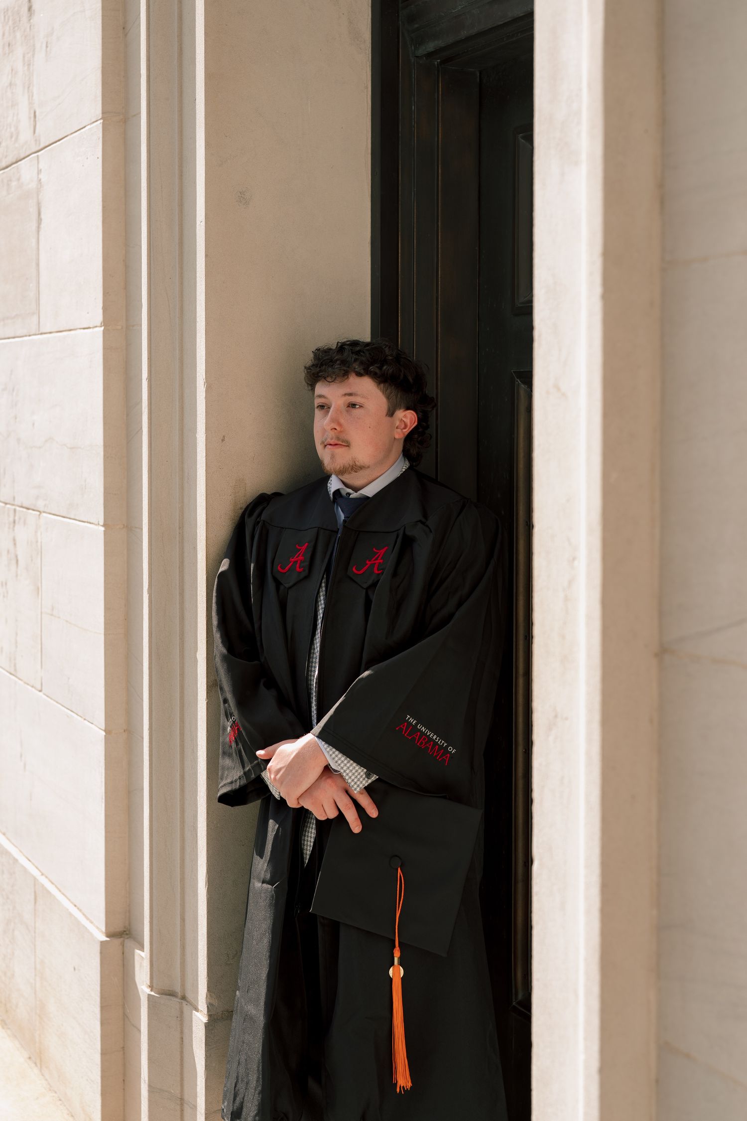 Young man in black graduation gown with red emblem stands in doorway, holding diploma with orange tassel.
