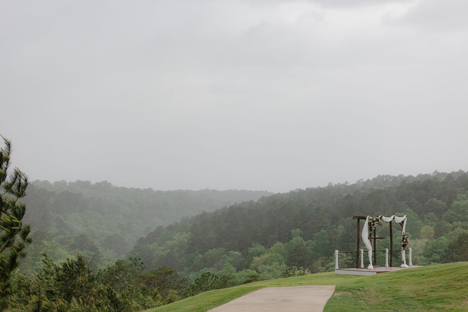 Misty outdoor wedding ceremony setup on a hilltop overlooking a vast green forested valley on a cloudy day.