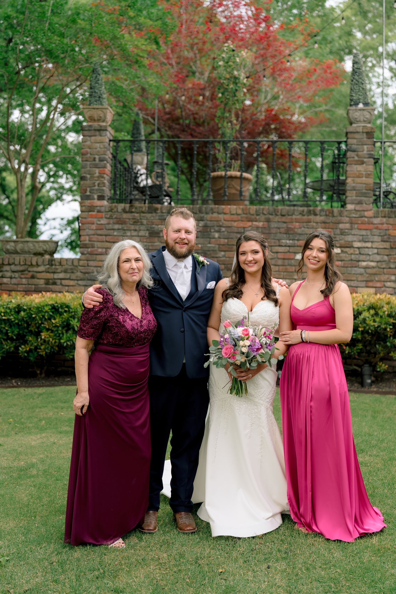 Wedding party group photo with bride, groom, and two women in burgundy and pink gowns outdoors.