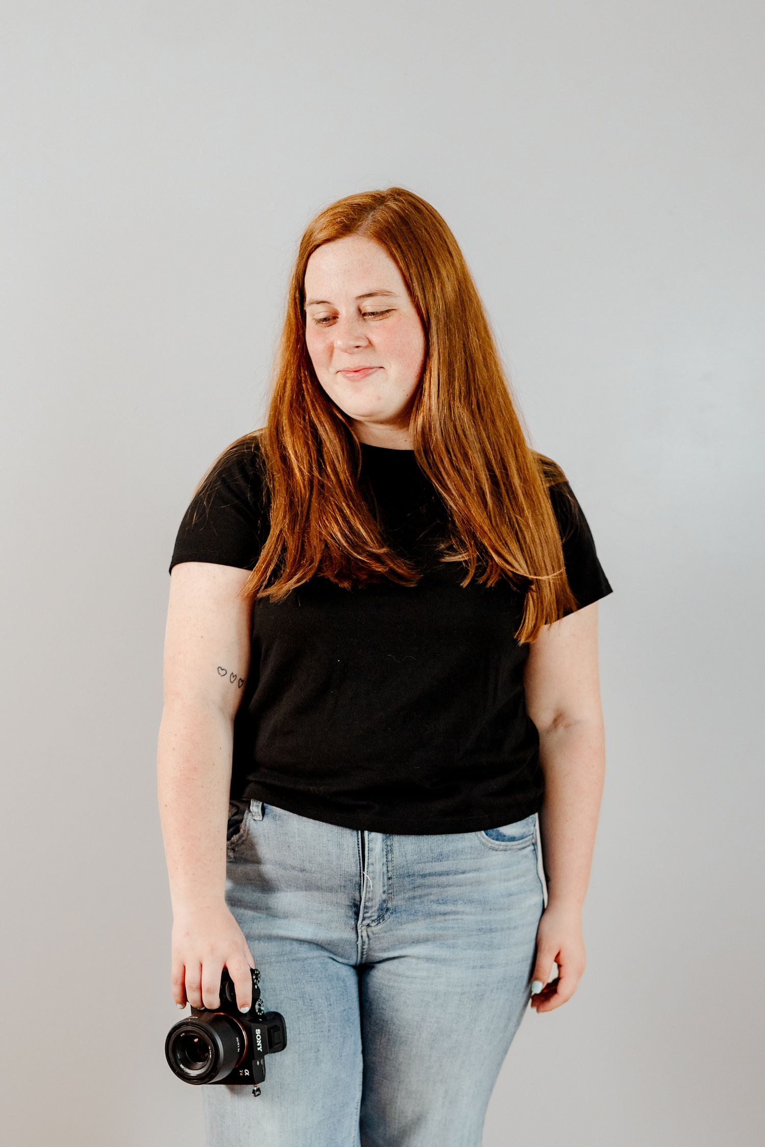 Person in casual attire poses with camera equipment against minimalist grey background.