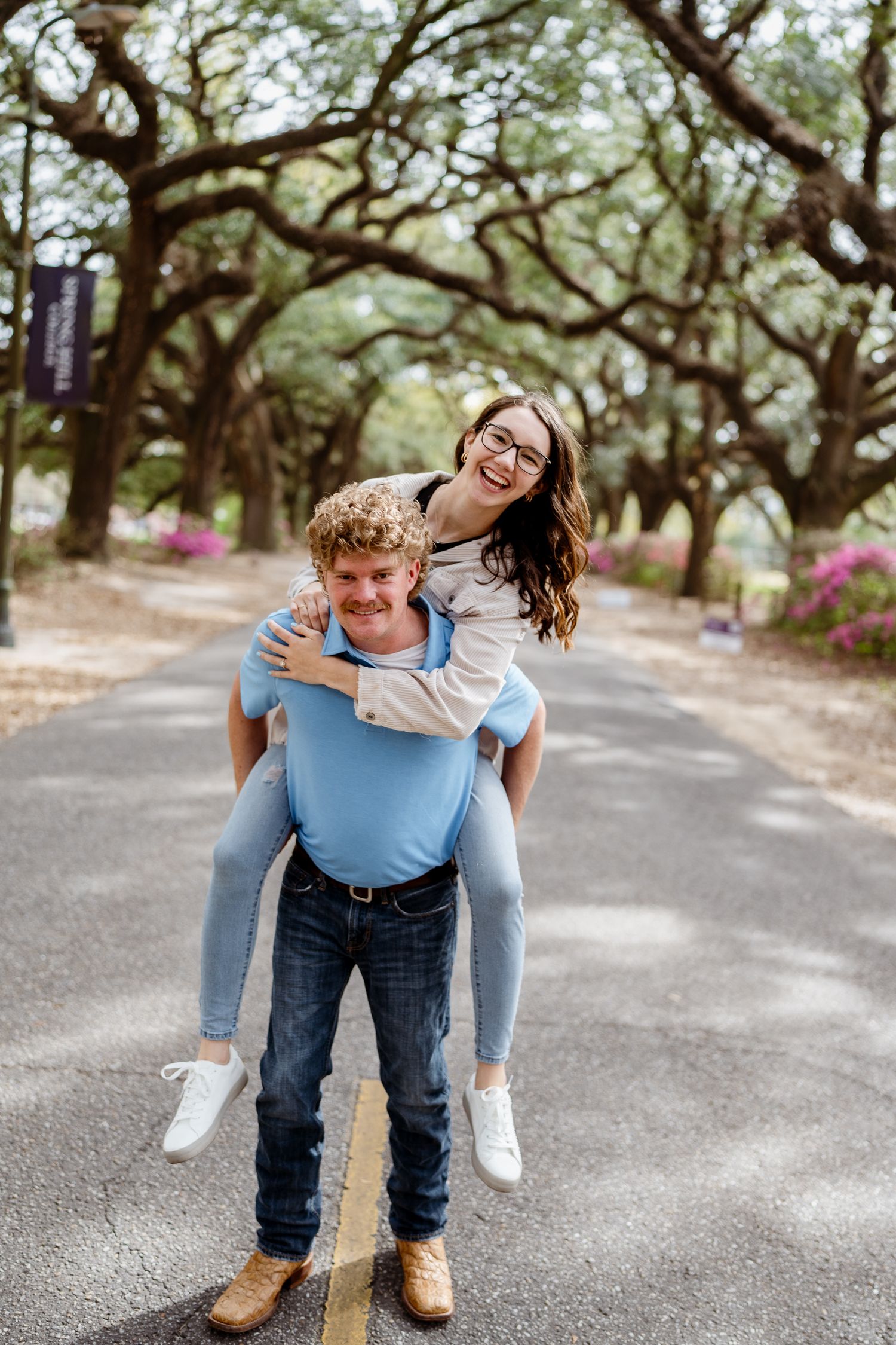 Two people playfully giving piggyback rides on a path under oak trees.