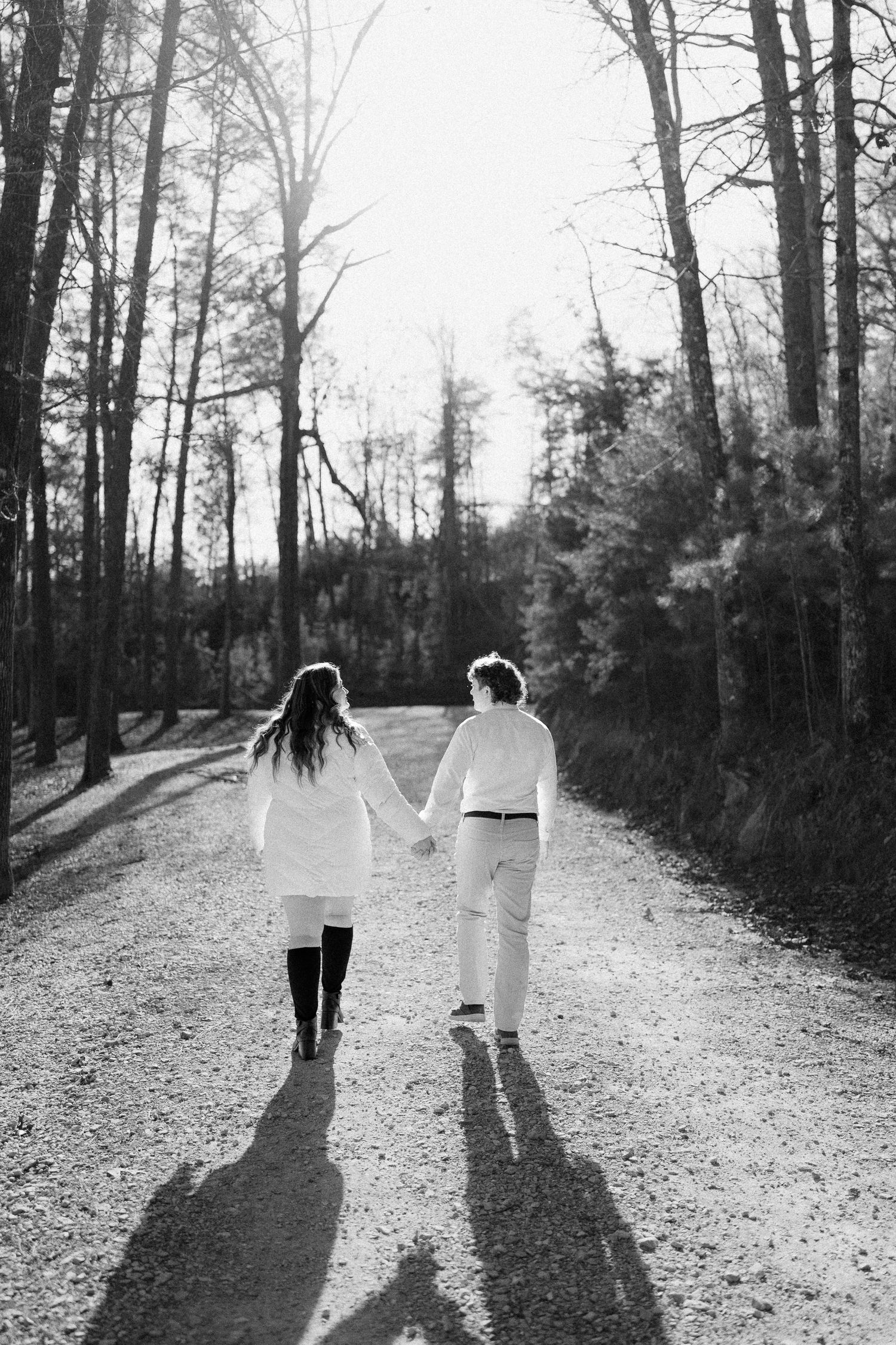 Black and white photo of couple walking hand in hand down a sunlit forest path.