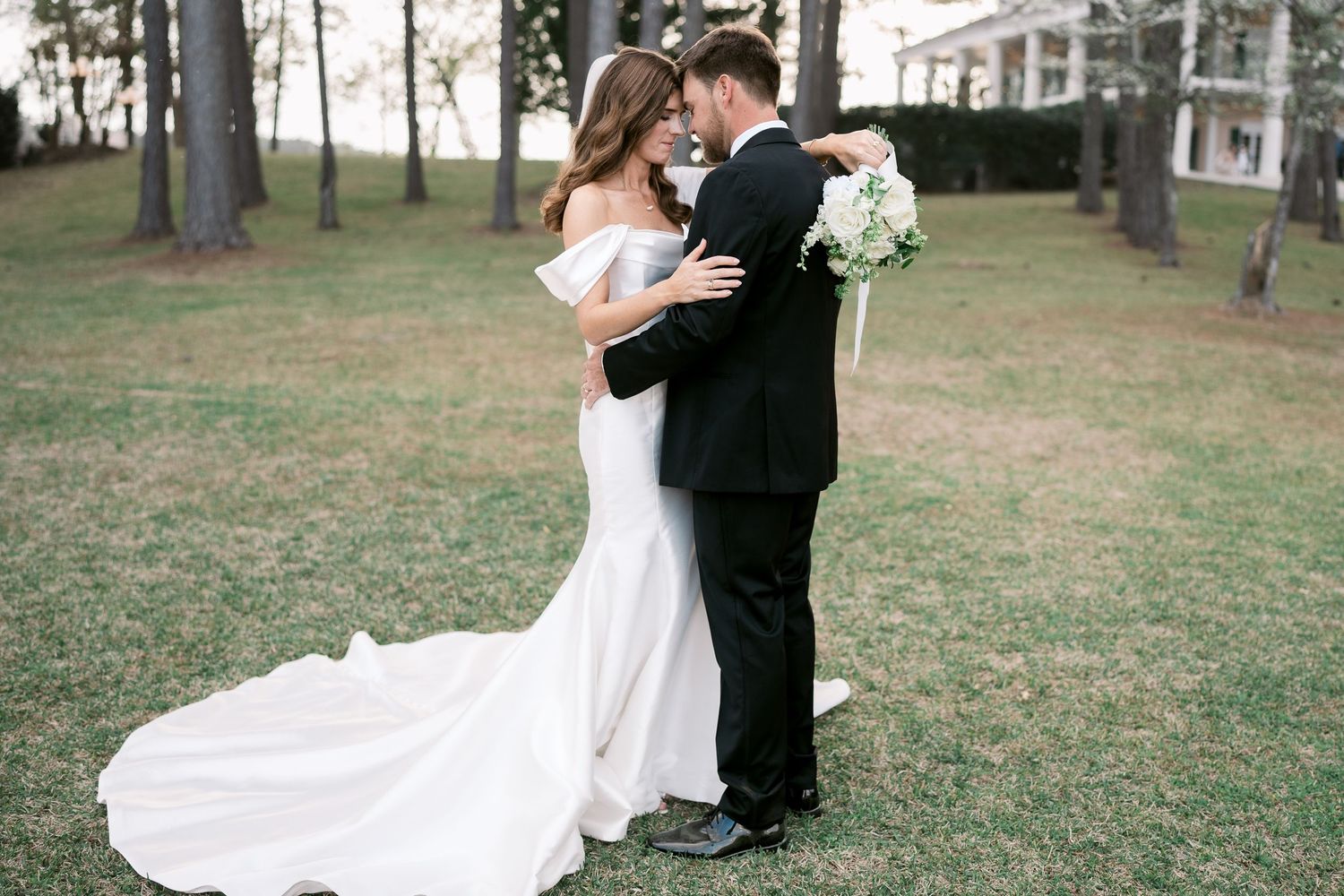 Elegant couple shares romantic moment in white gown and black tuxedo on lawn.