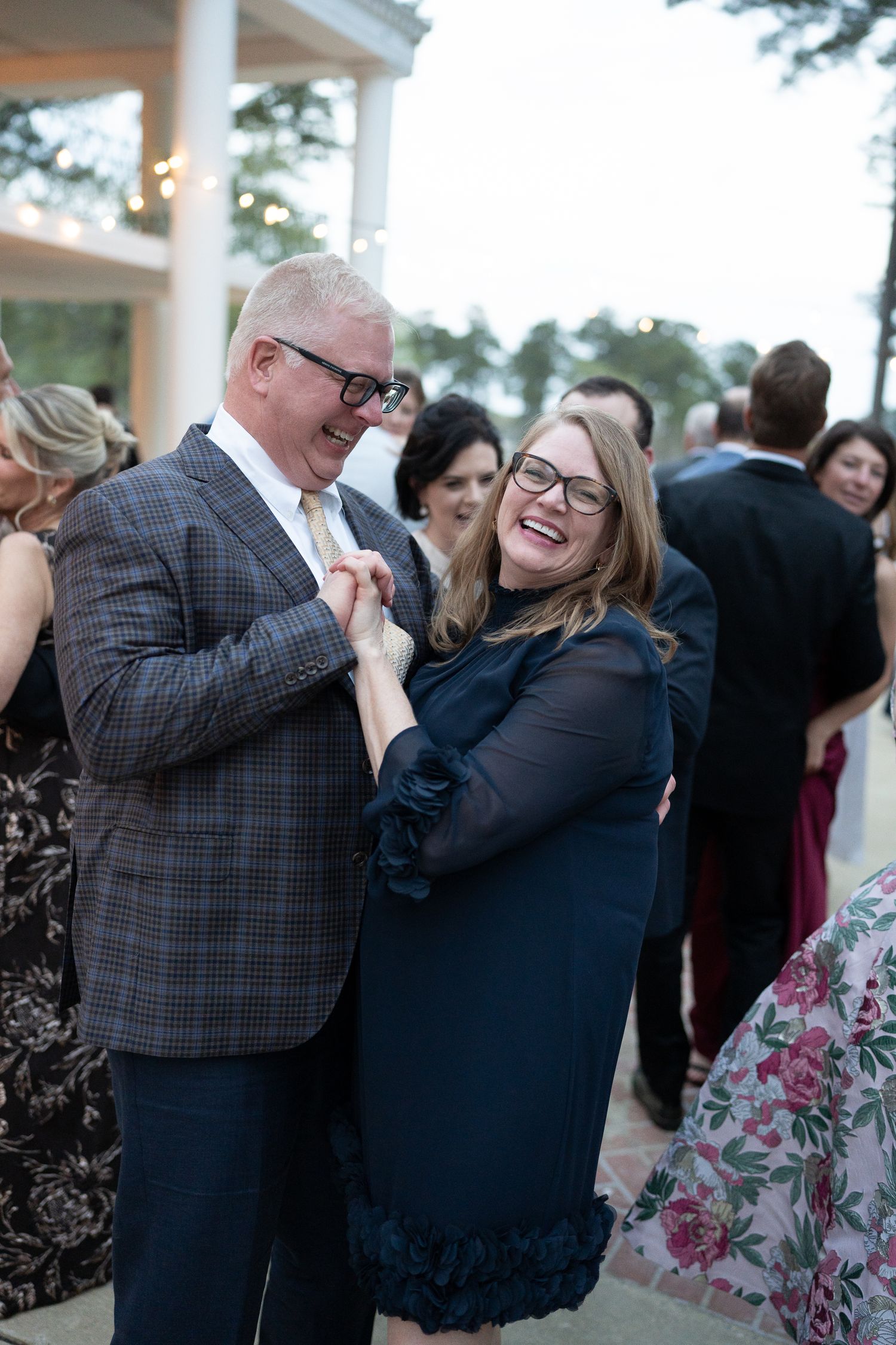 Wedding guests dance and embrace at an outdoor reception under a covered patio area.
