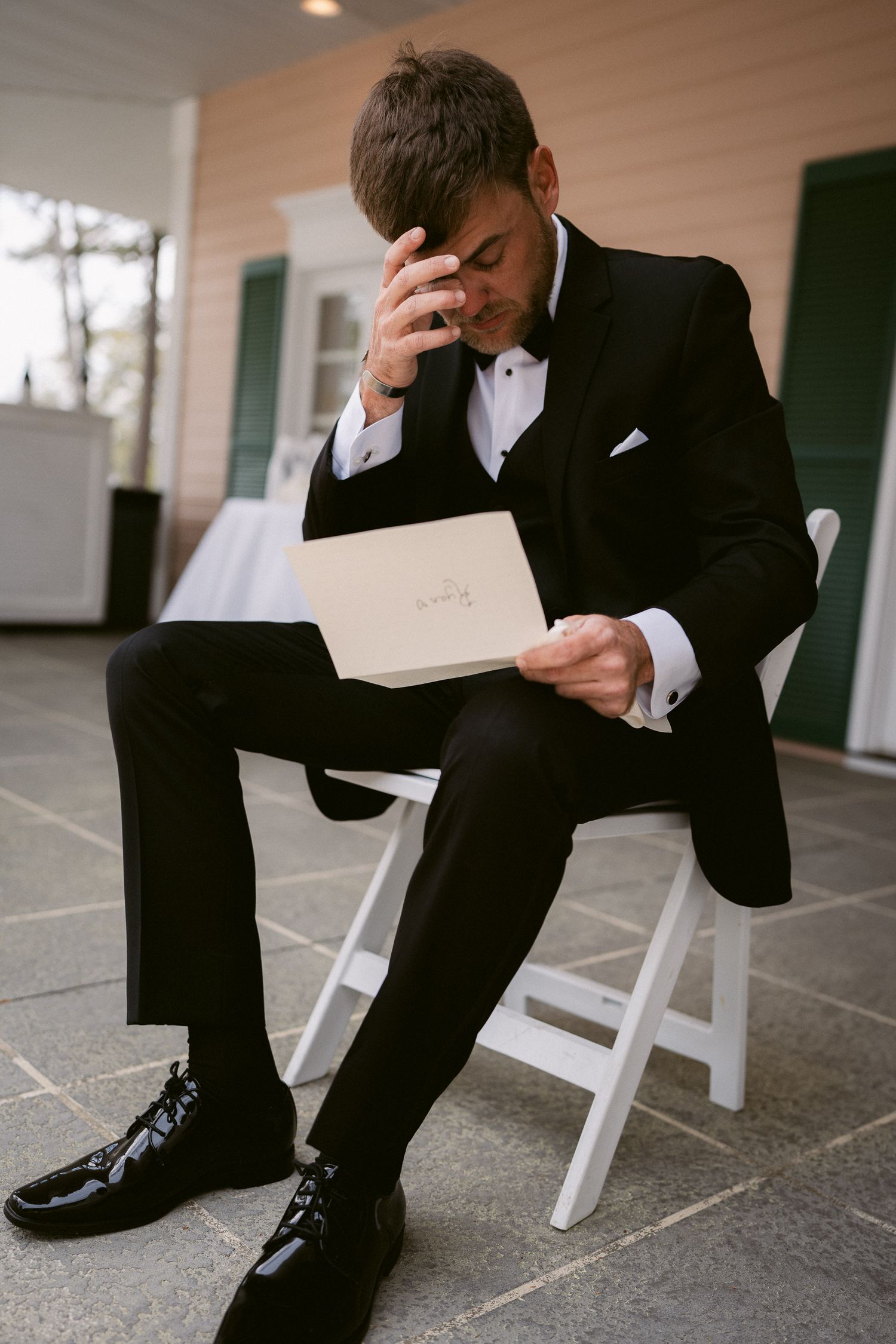 Groom in classic tuxedo sits reading an emotional letter on his wedding day.