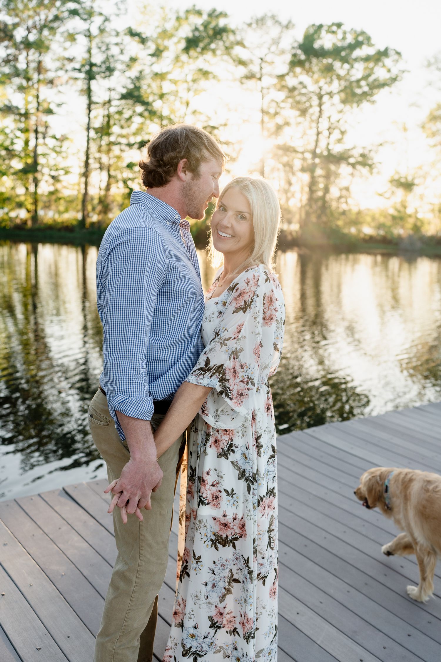 A couple shares an intimate moment on a dock while their dog walks in the background.