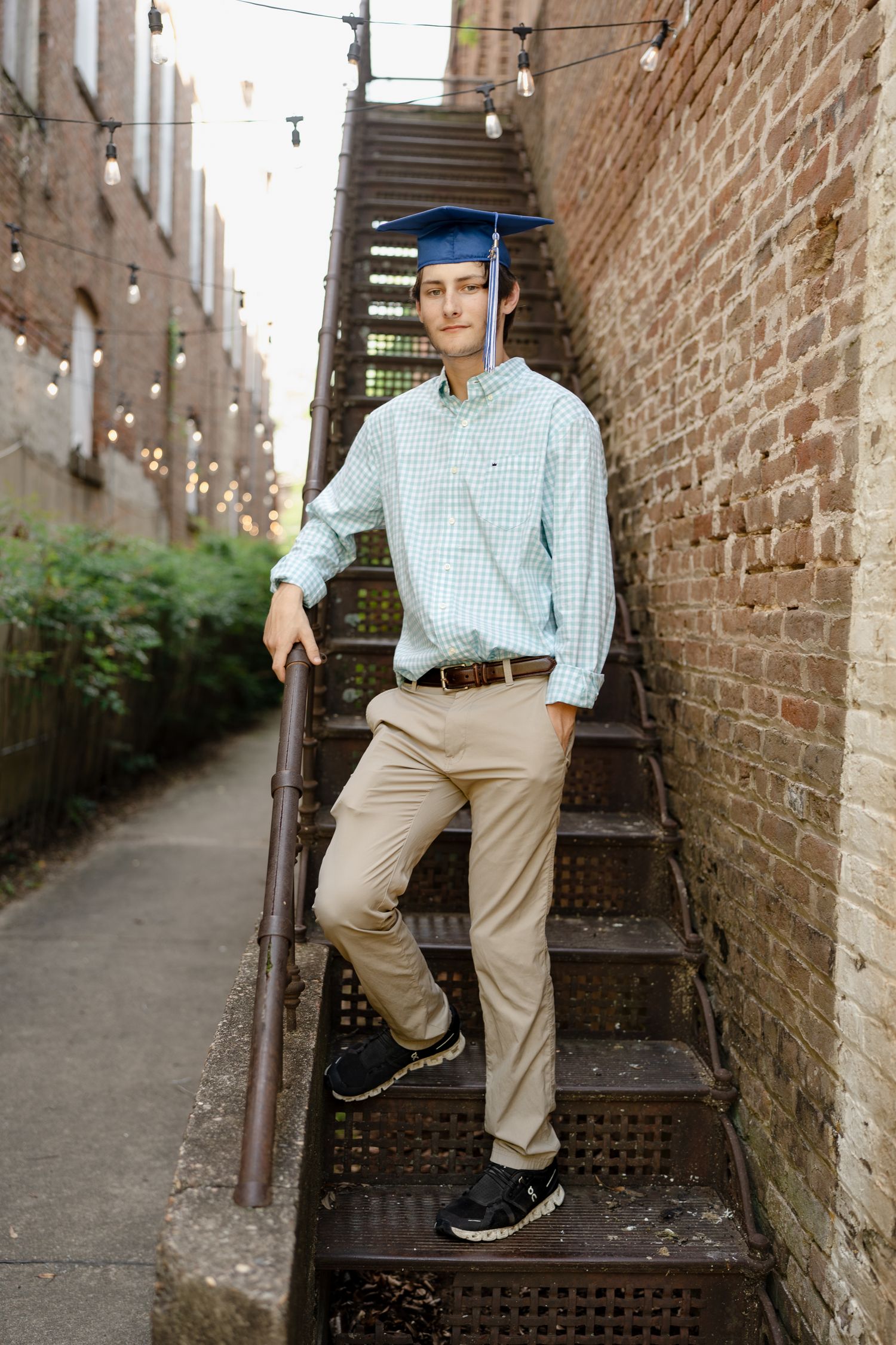 Graduate in casual attire poses on metal staircase between brick walls with string lights.