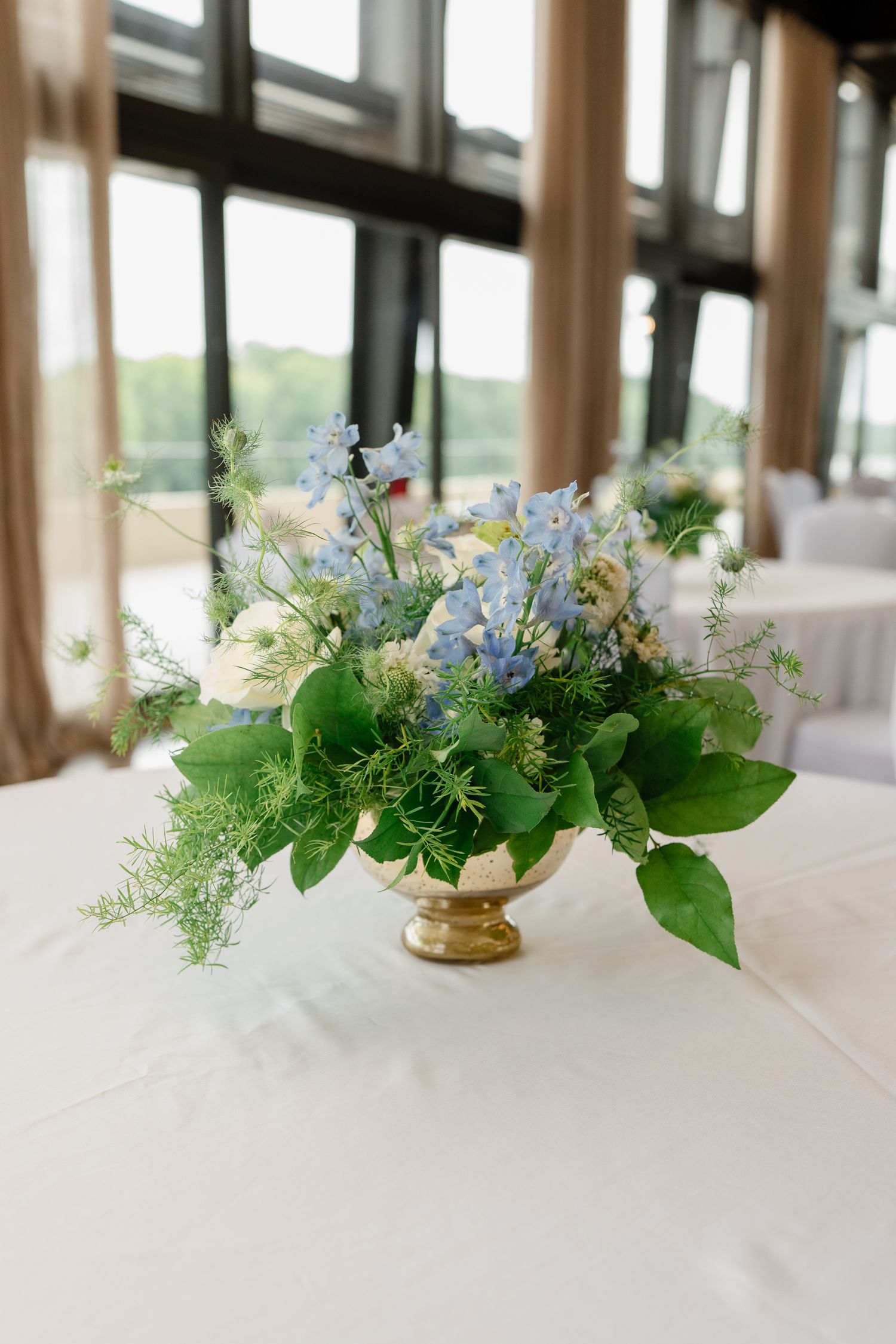Elegant floral centerpiece with white and light blue flowers in a gold pedestal vase on a white tablecloth.
